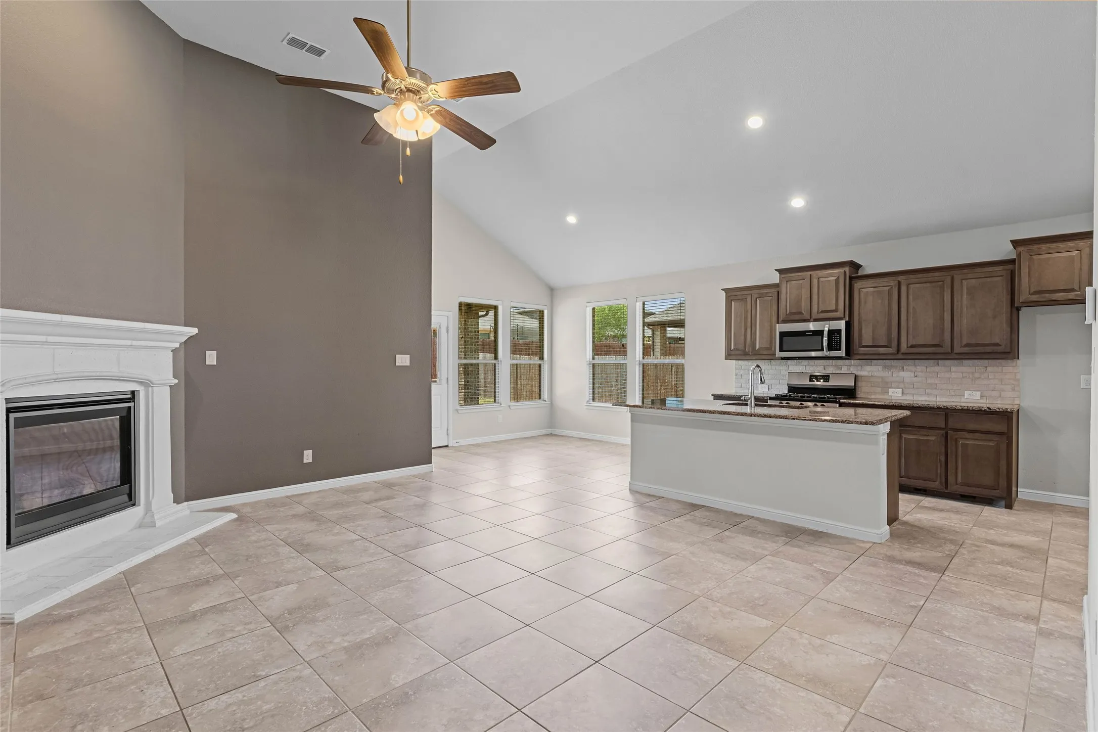 Vaulted ceiling with a ceiling fan, fireplace with a white mantel, tiled flooring, kitchen island with a sink, and dark wood-finish cabinetry