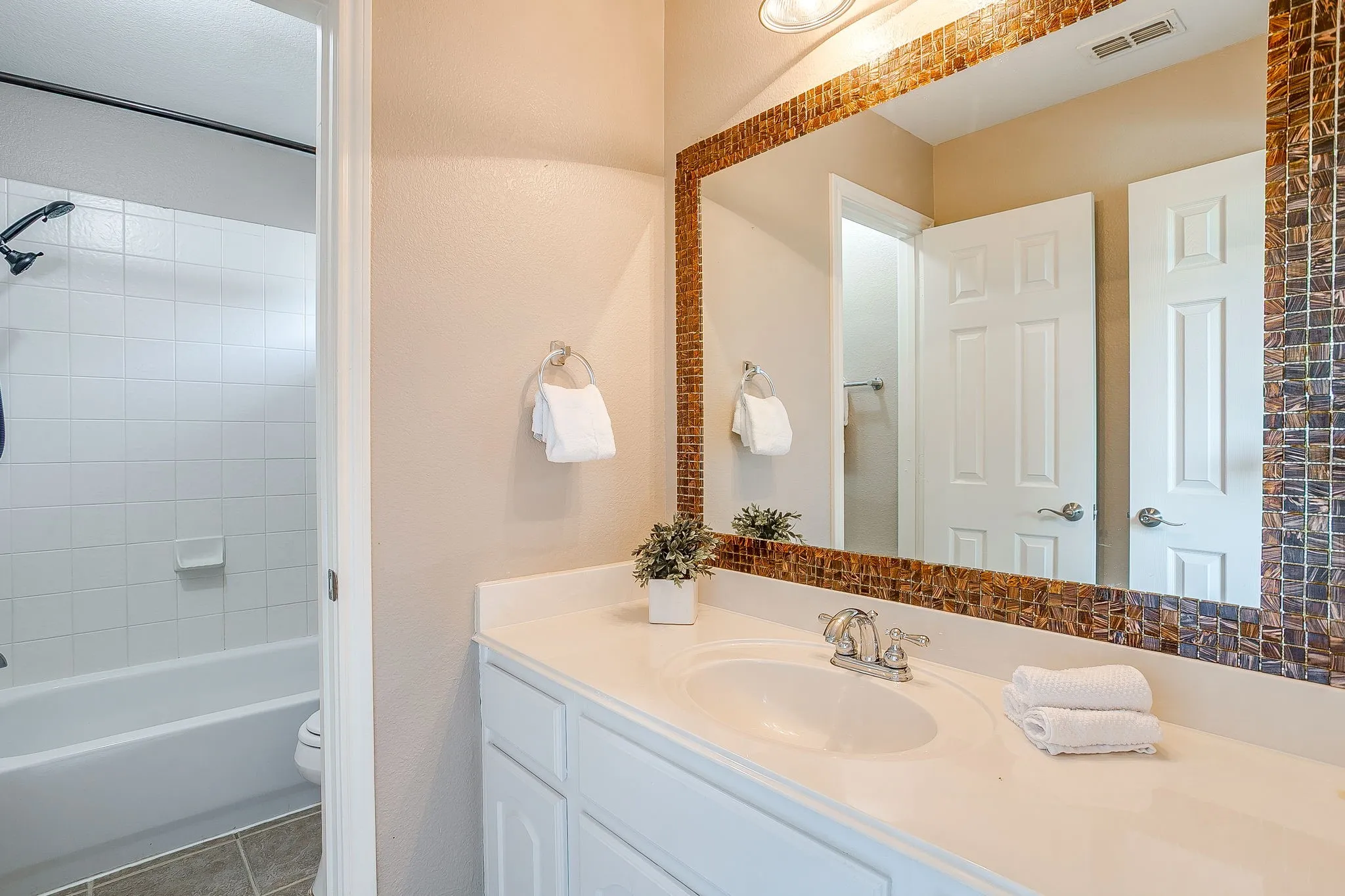 White vanity with an integrated sink and chrome faucet, mosaic-tiled mirror frame, and a white tiled shower and tub combination