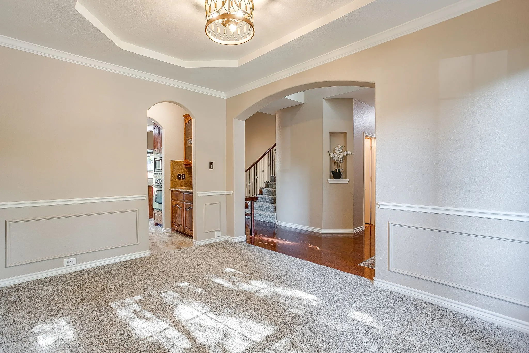 Tray ceiling with decorative lighting fixture, carpeted flooring, wainscoting paneling, arched doorways, and wood-finish flooring in adjacent areas