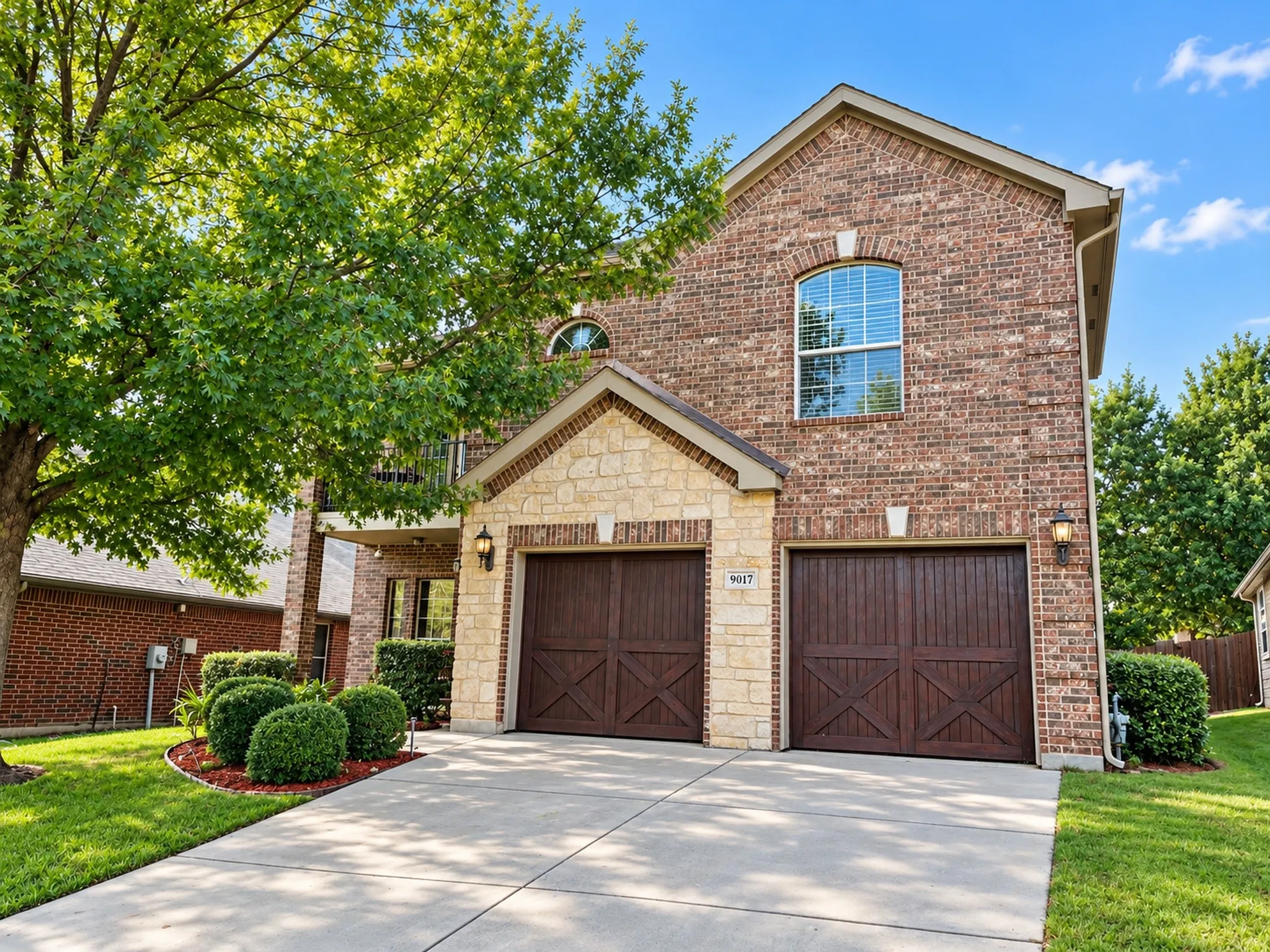 Brick and stone exterior with arched windows, two wood-finish garage doors, concrete driveway, and manicured landscaping
