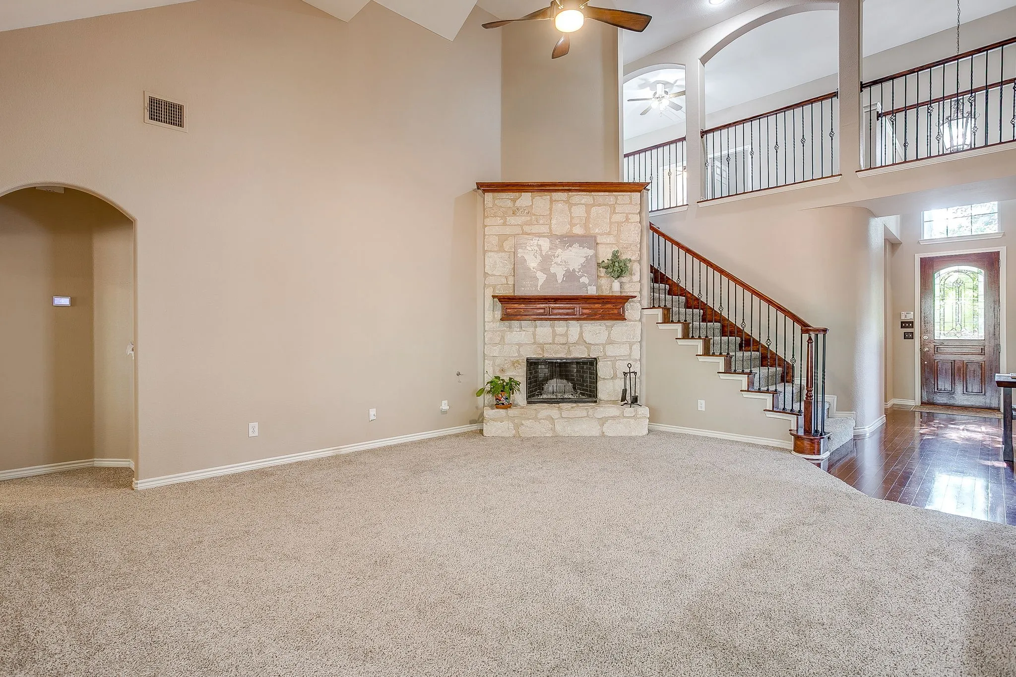 Vaulted ceiling with ceiling fan, stone fireplace with wood mantel, staircase with dark wood handrail and wrought iron balusters, arched entryway, and wood-finish flooring in the entryway