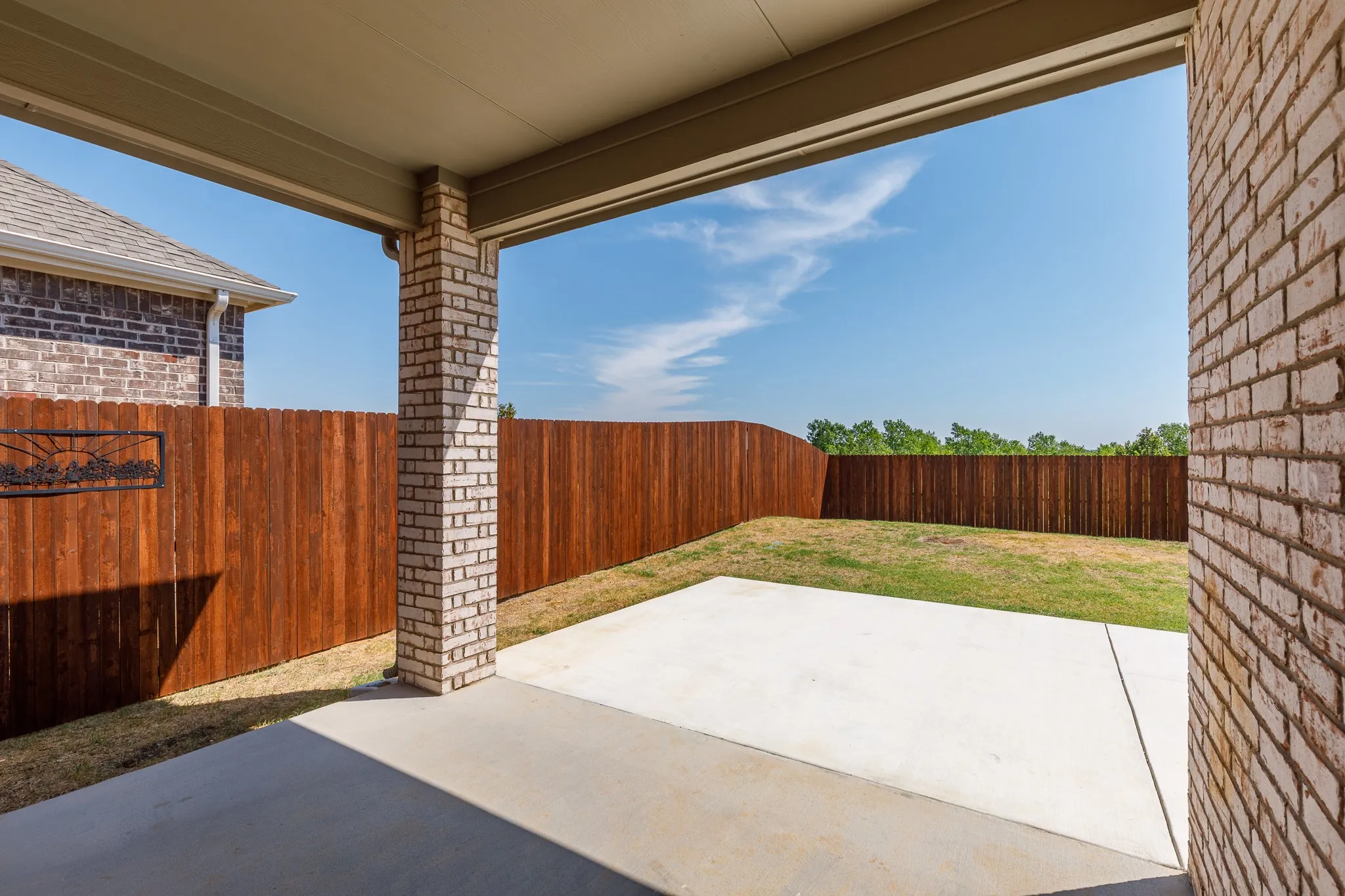 Covered patio with a concrete surface, supported by a light brick column, opening onto a fenced yard with grass and a wooden privacy fence