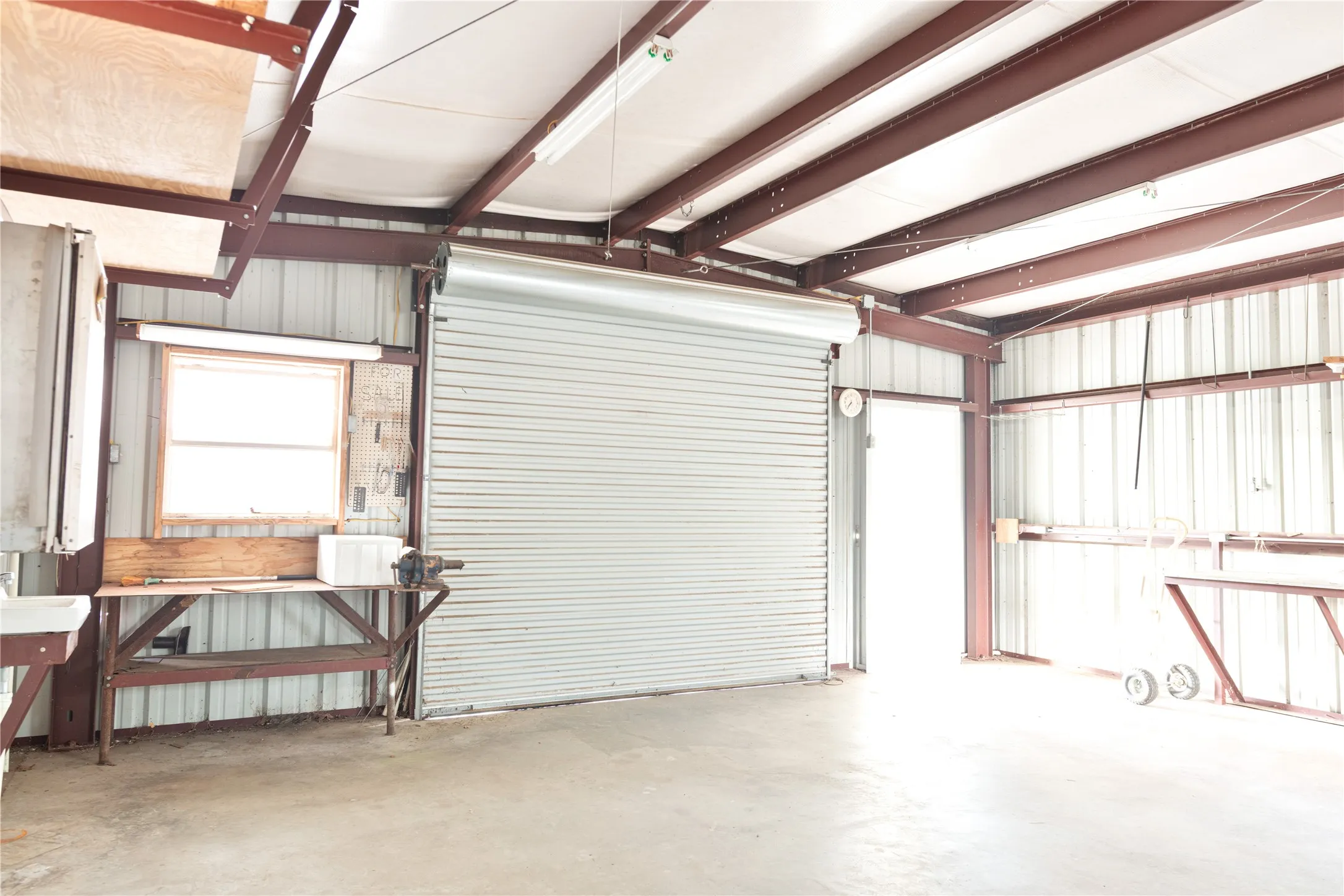 Concrete flooring, exposed metal beam ceiling, corrugated metal walls, roll-up door, and workbench with window