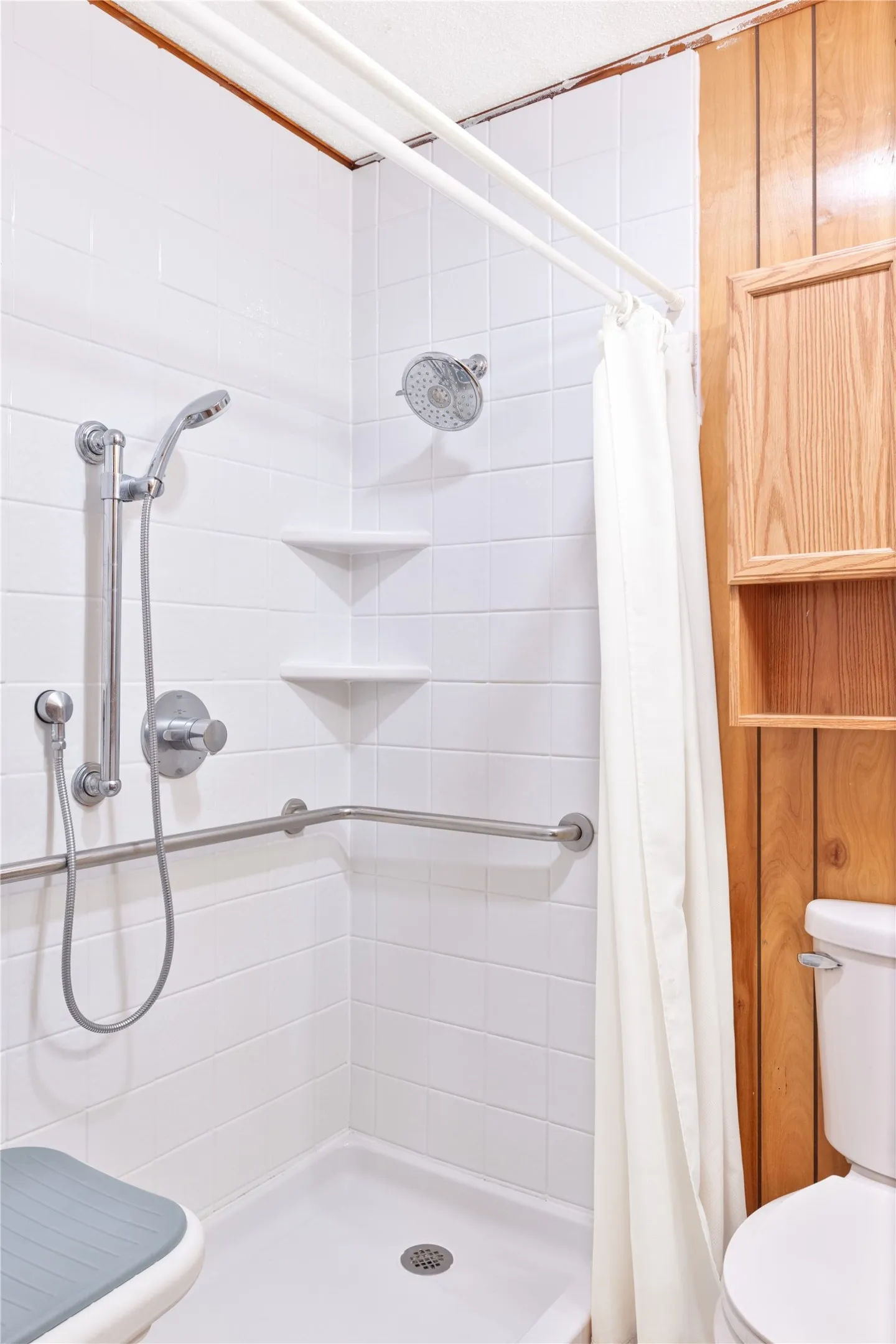 Shower stall with white tile surround, a built-in corner shelf, and grab bar