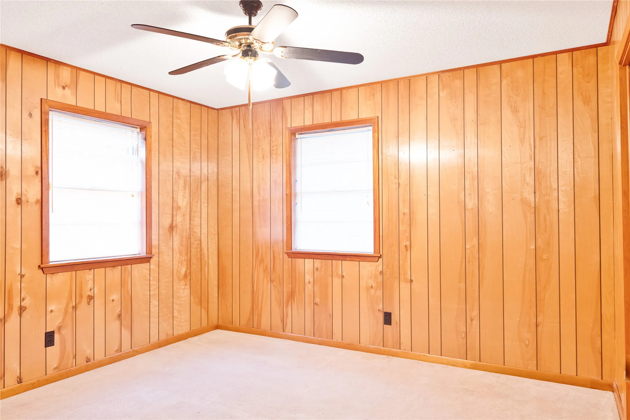 Wood paneling on walls, two window openings, ceiling fan with integrated lighting, and carpeted flooring