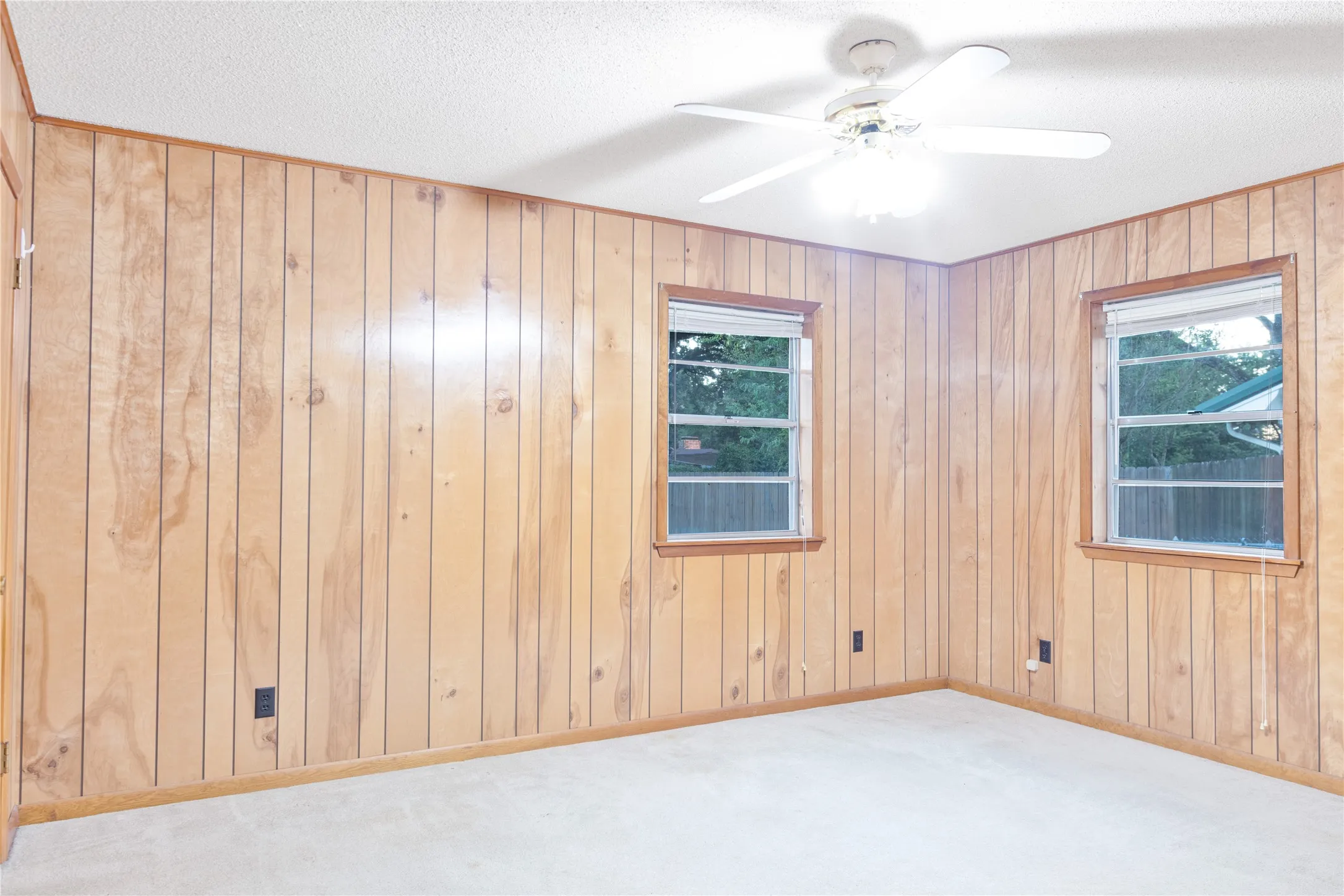 Wood paneling, two windows, textured ceiling, ceiling fan with integrated lighting, and light-toned carpeting