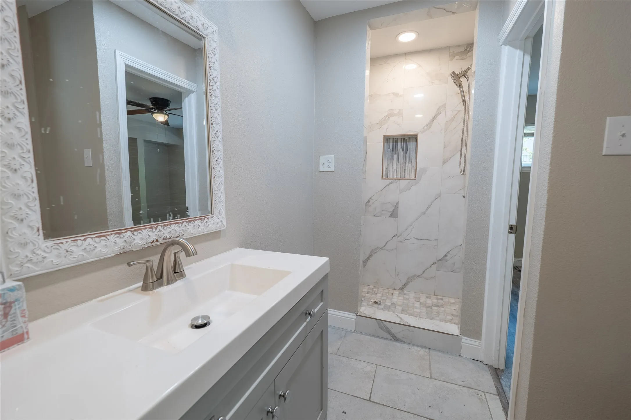 Vanity with integrated sink, brushed nickel faucet, decorative framed mirror, walk-in shower with marble-look tile, and recessed shower niche