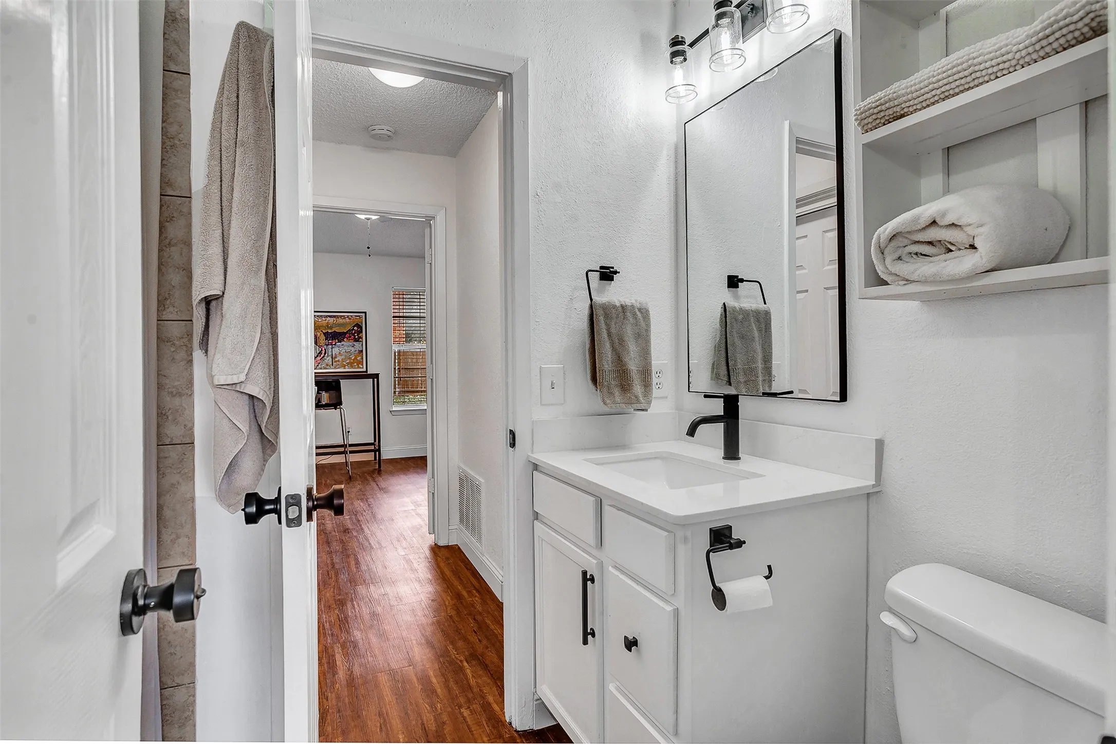 White vanity with a single basin sink and matte black faucet, framed mirror with overhead lighting, white built-in shelving, white toilet, and wood-finish flooring