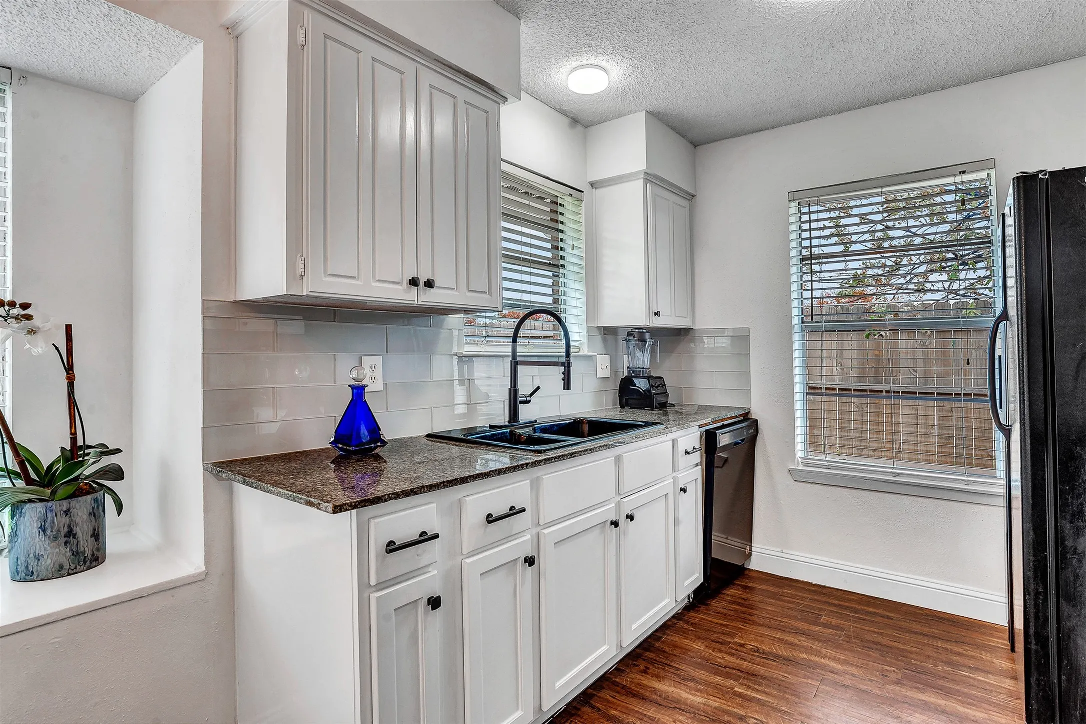 Kitchen featuring white shaker-style cabinetry, dark granite countertops, a subway tile backsplash, a black double-basin sink with a gooseneck faucet, and wood-finish flooring