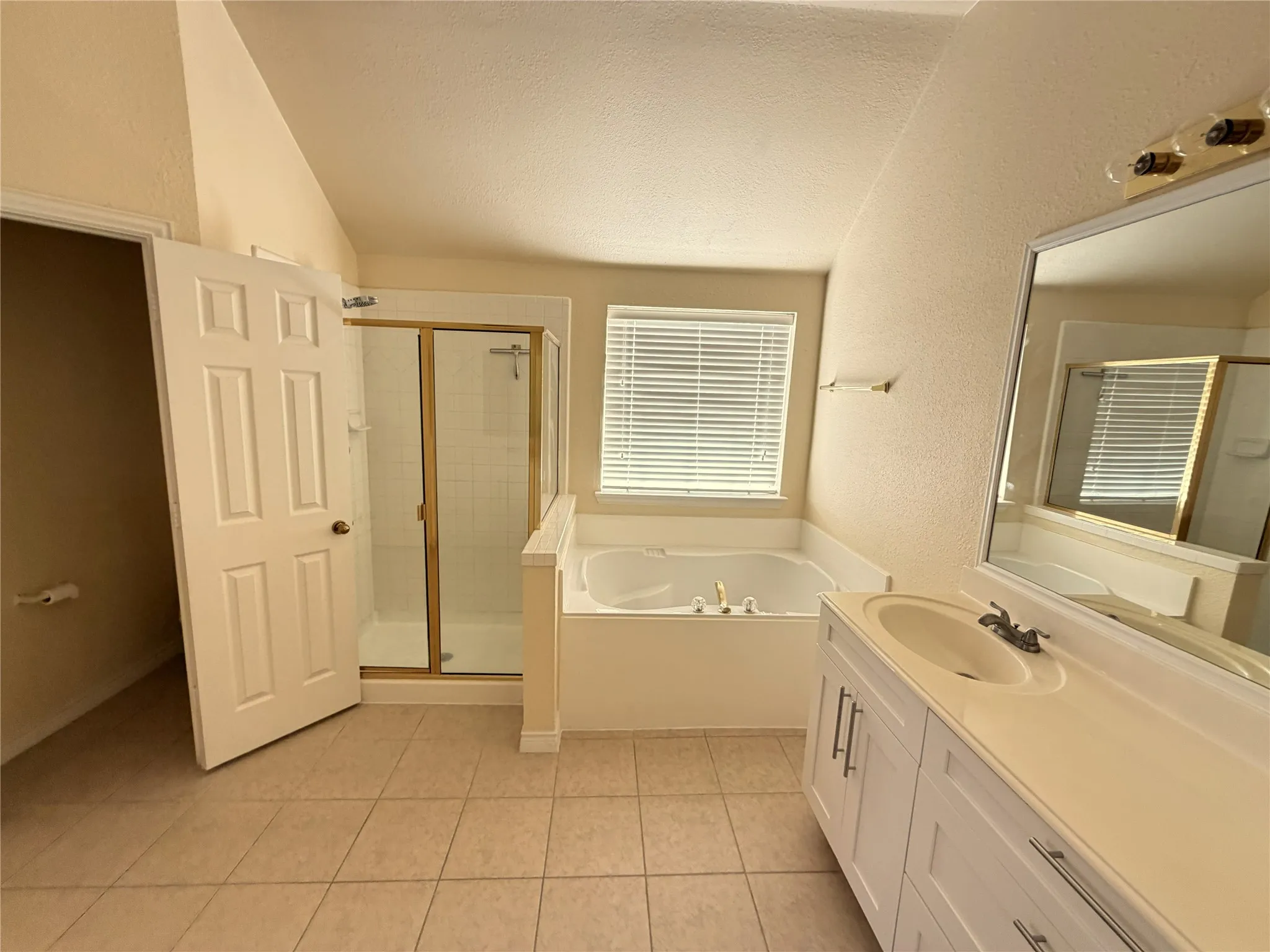 Tiled flooring, white vanity with dual sinks, white countertop, built-in bathtub, and enclosed shower with gold-toned trim
