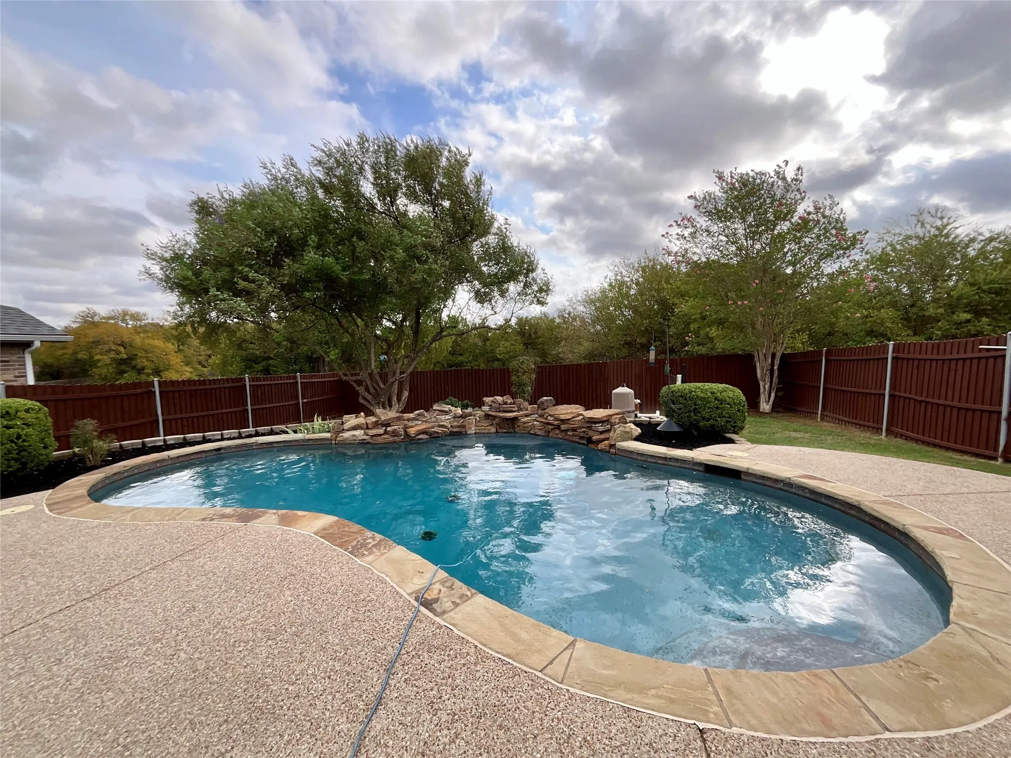 Curved swimming pool with natural stone coping, expansive aggregate deck, rock waterfall feature, and privacy fencing