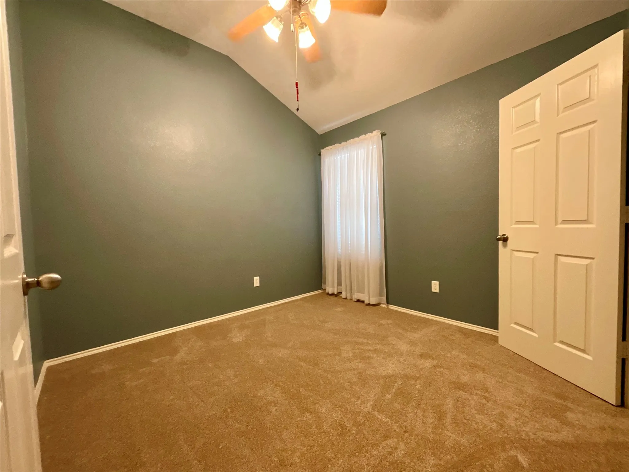 Vaulted ceiling, carpeted flooring, paneled interior door, ceiling fan with light fixture, window with sheer curtains