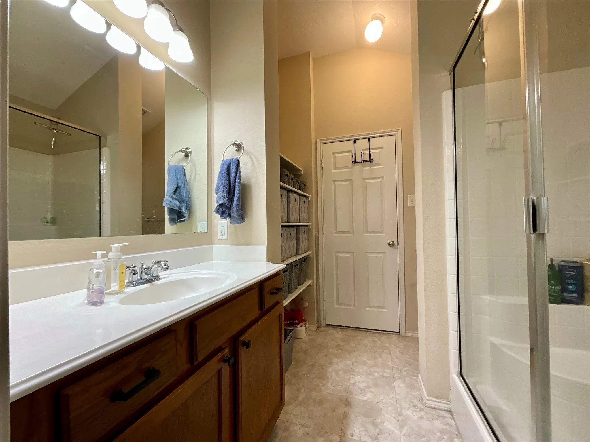 Single vanity with integrated sink and chrome faucet, wood-finish cabinetry, built-in shelving, and a glass-enclosed shower