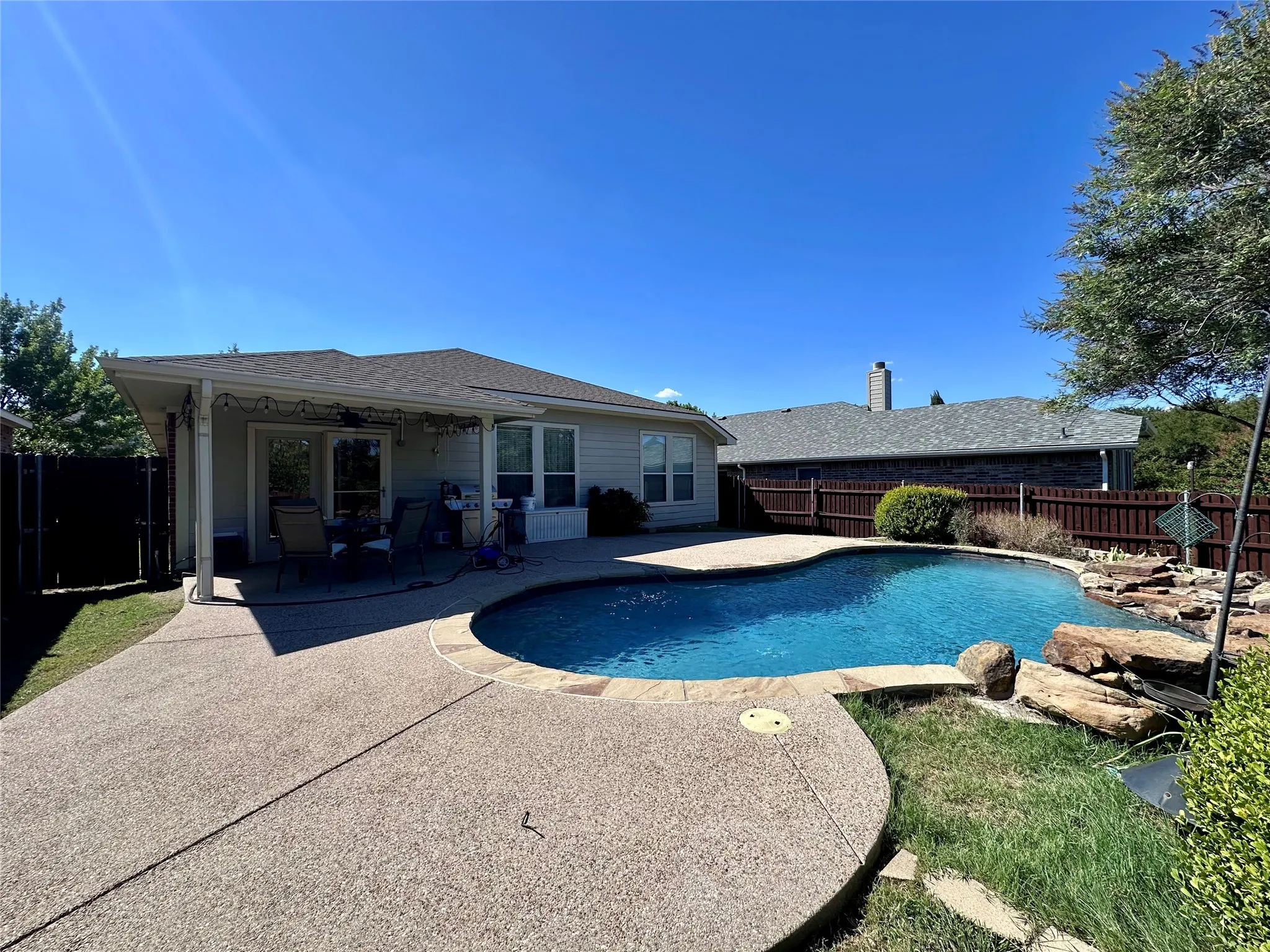 Backyard oasis featuring a freeform swimming pool with rock detailing, extensive concrete decking, and a covered patio area