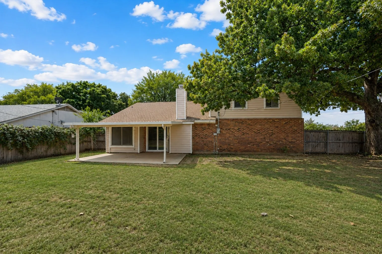 Expansive green lawn, covered patio, brick and siding exterior, privacy fencing, and mature trees