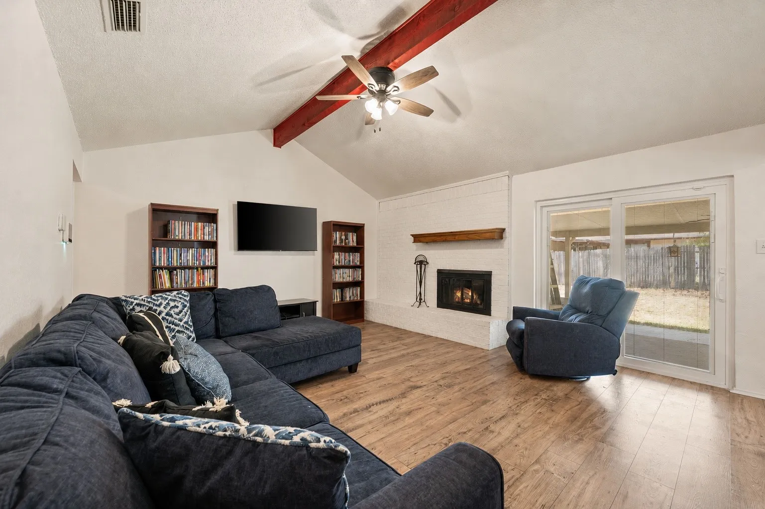 Spacious vaulted ceiling living area featuring wood-finish flooring, built-in shelving with display cabinetry, wall-mounted flat-screen television, gas fireplace with surround, and red ceiling fan with integrated lighting