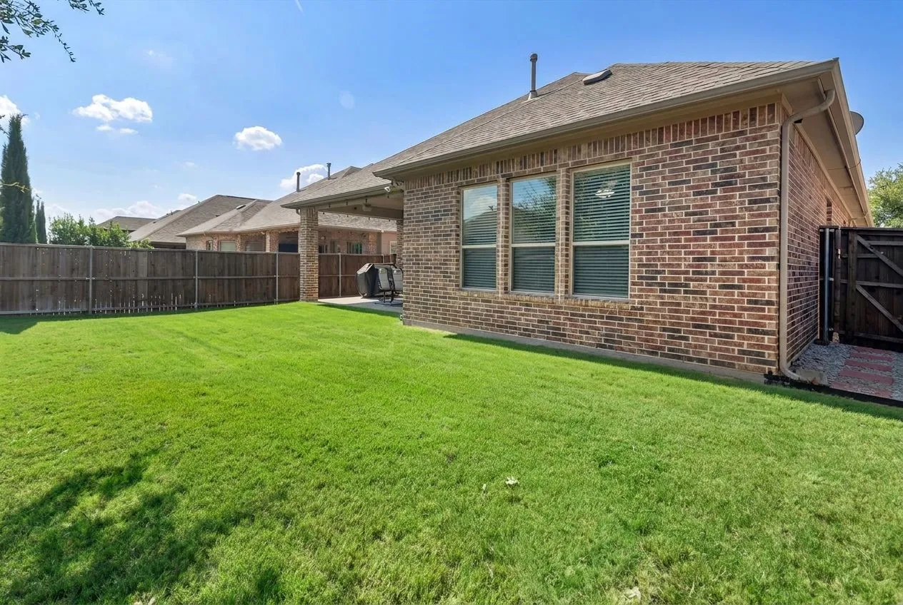 Expansive backyard featuring a manicured lawn, brick exterior, covered patio, and wood privacy fencing