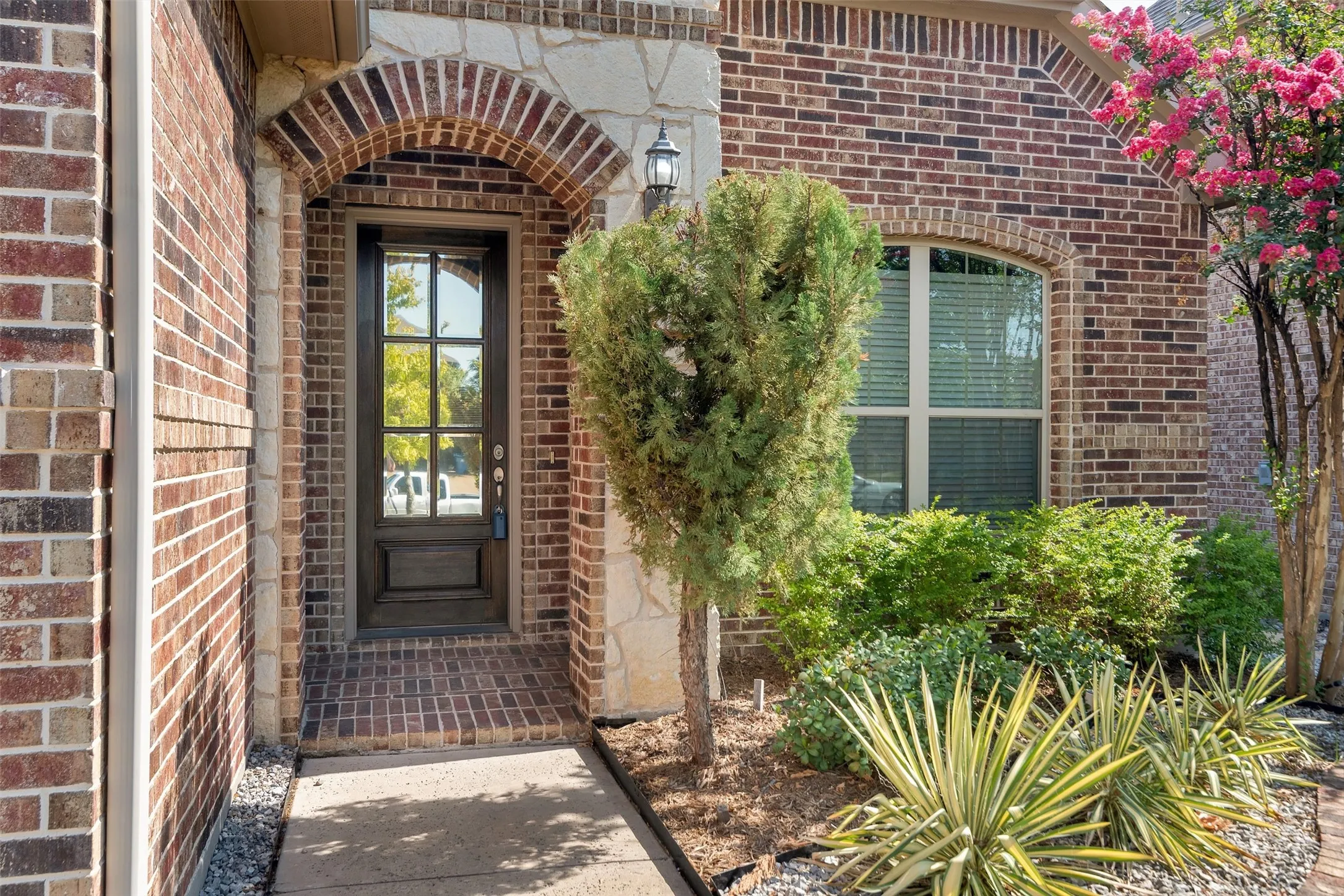 Brick exterior with stone accents, arched entryway, dark wood-finish door with glass panels, and a well-maintained planting bed