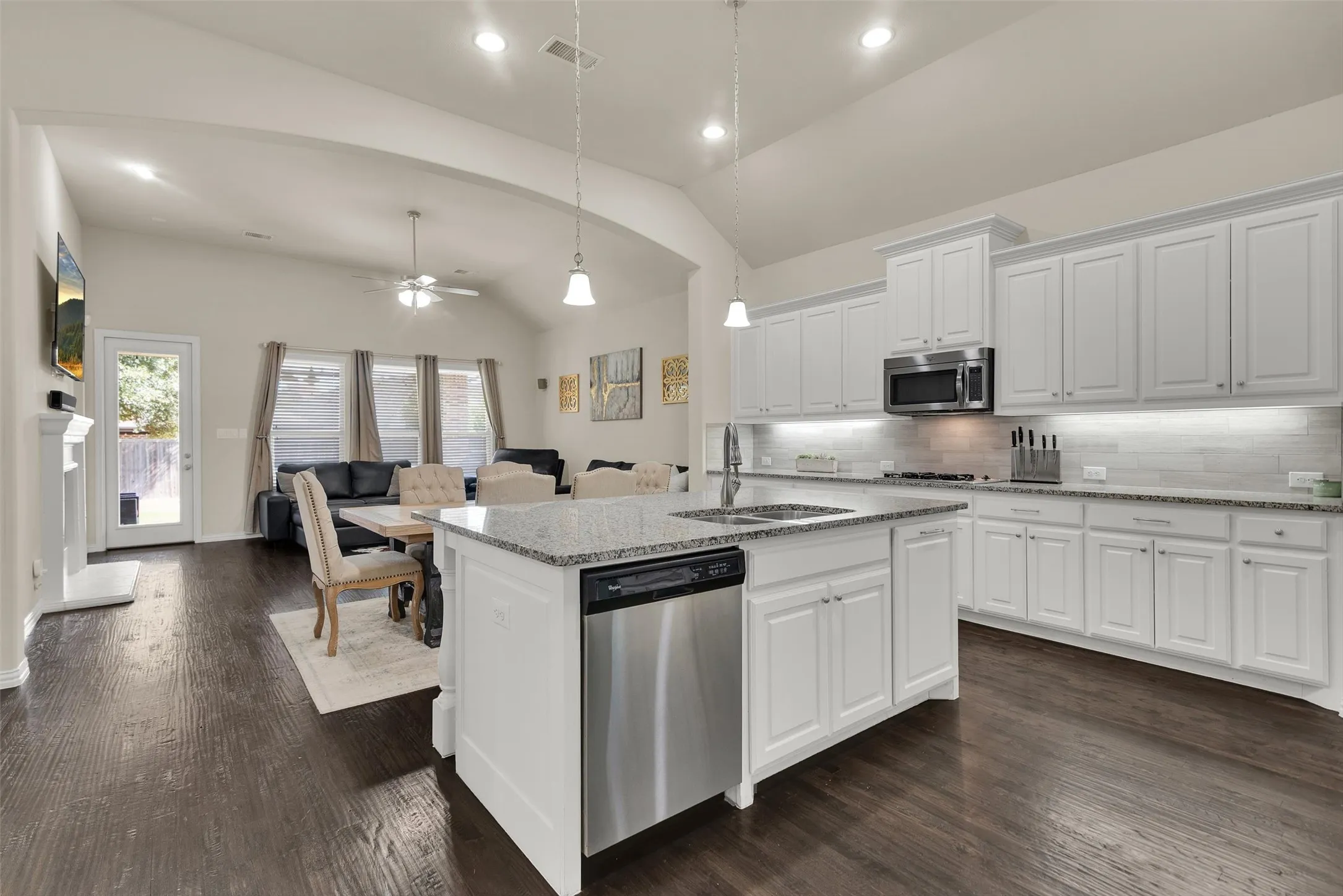Open-concept living space with vaulted ceilings, wood-finish flooring, a kitchen island featuring a sink and dishwasher, white cabinetry, and a subway tile backsplash
