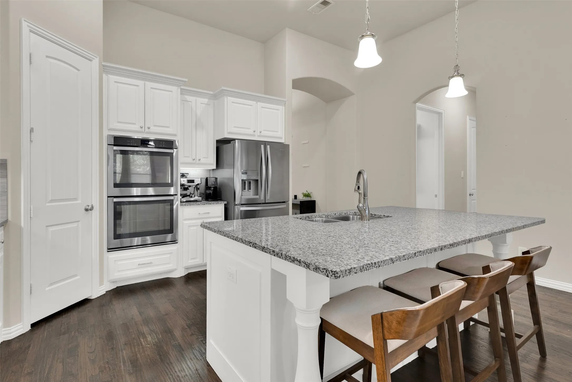 Kitchen featuring white cabinetry, stainless steel double ovens, a large center island with a sink, pendant lighting, and dark wood-finish flooring