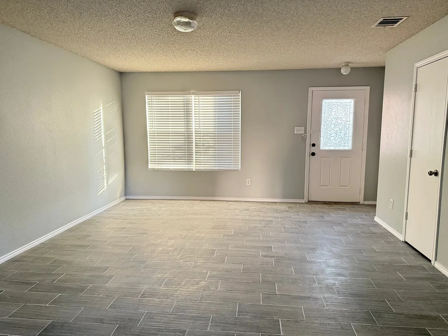 Gray tile-pattern flooring, window with blinds, paneled door with frosted glass, flush-mount ceiling light