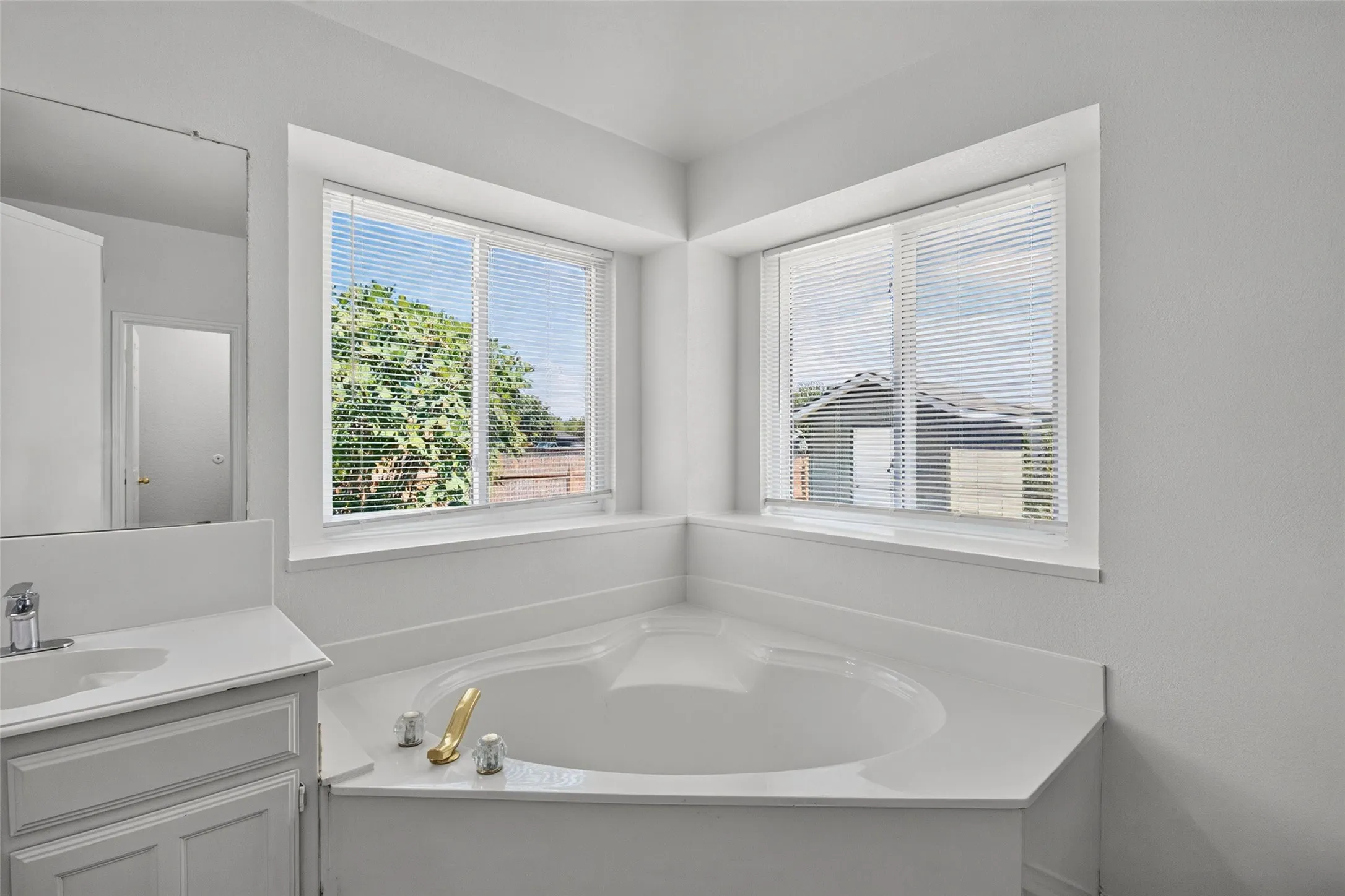 Corner soaking tub with integrated white surround, dual windows with blinds, and a white vanity with an integrated sink and chrome faucet