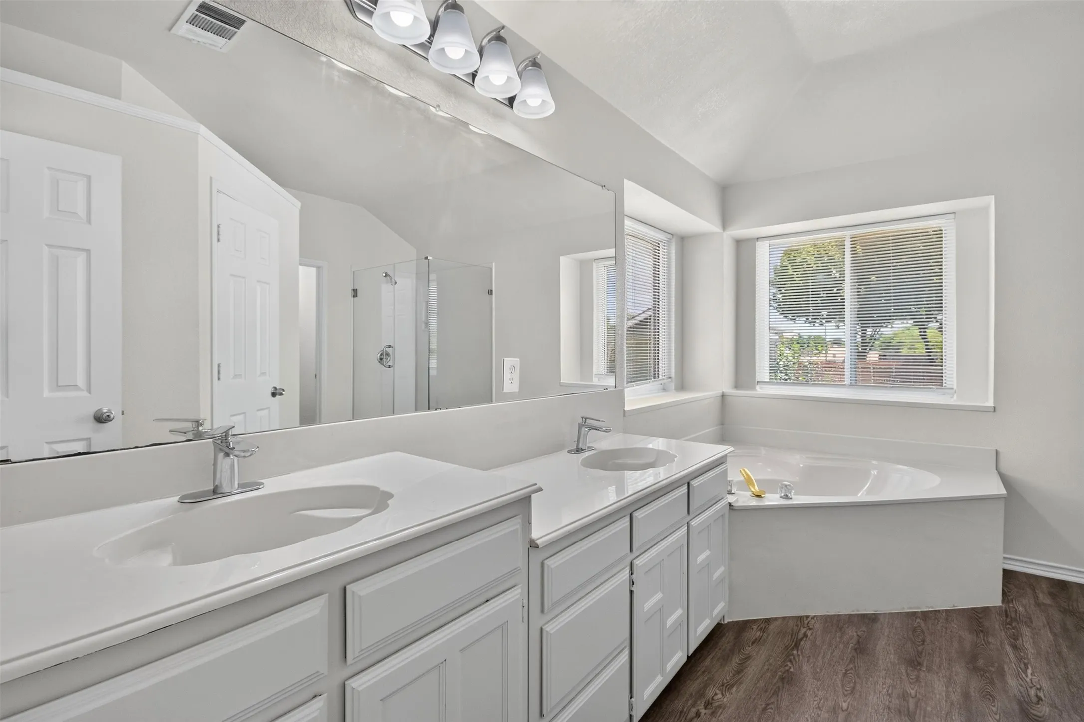 Dual vanity with white cabinetry and integrated sinks, wide-set mirror, corner soaking tub, and wood-finish flooring