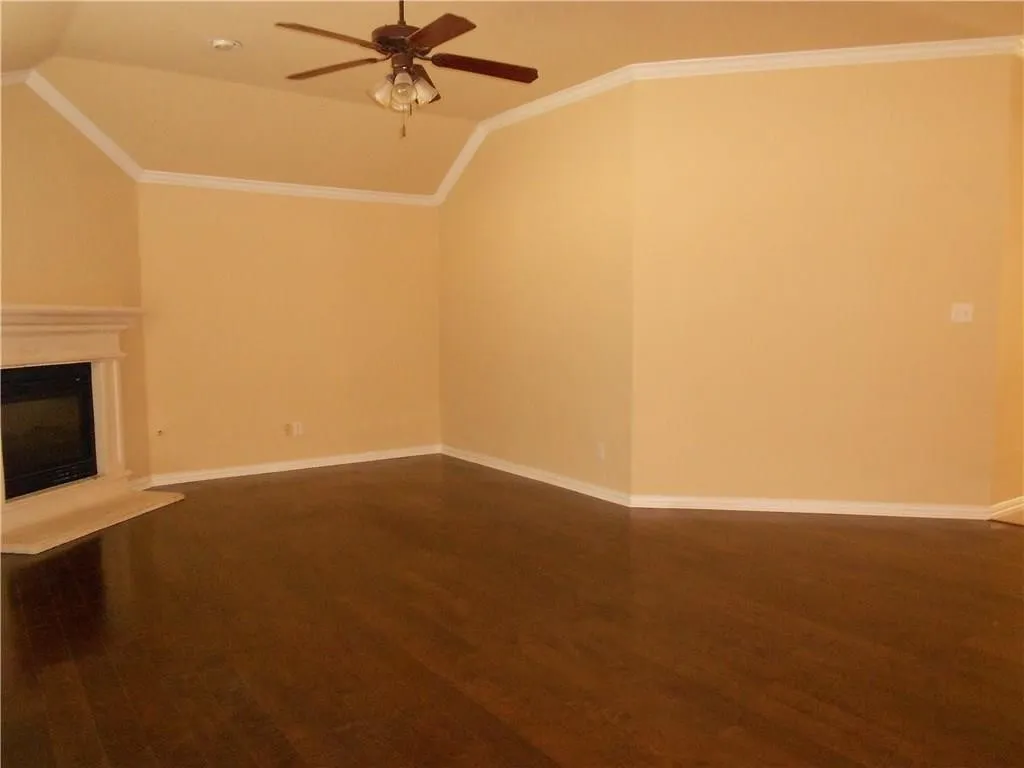 Vaulted ceiling with crown molding, wood-finish flooring, ceiling fan, and fireplace with a stone mantel