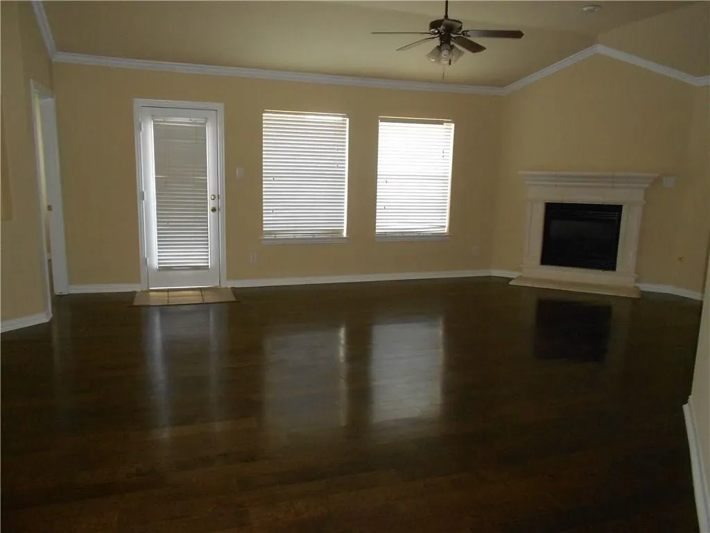 Spacious living area showcasing rich wood-finish flooring, abundant natural light from multiple windows and glass door entry, ceiling fan, and a fireplace with surround framing
