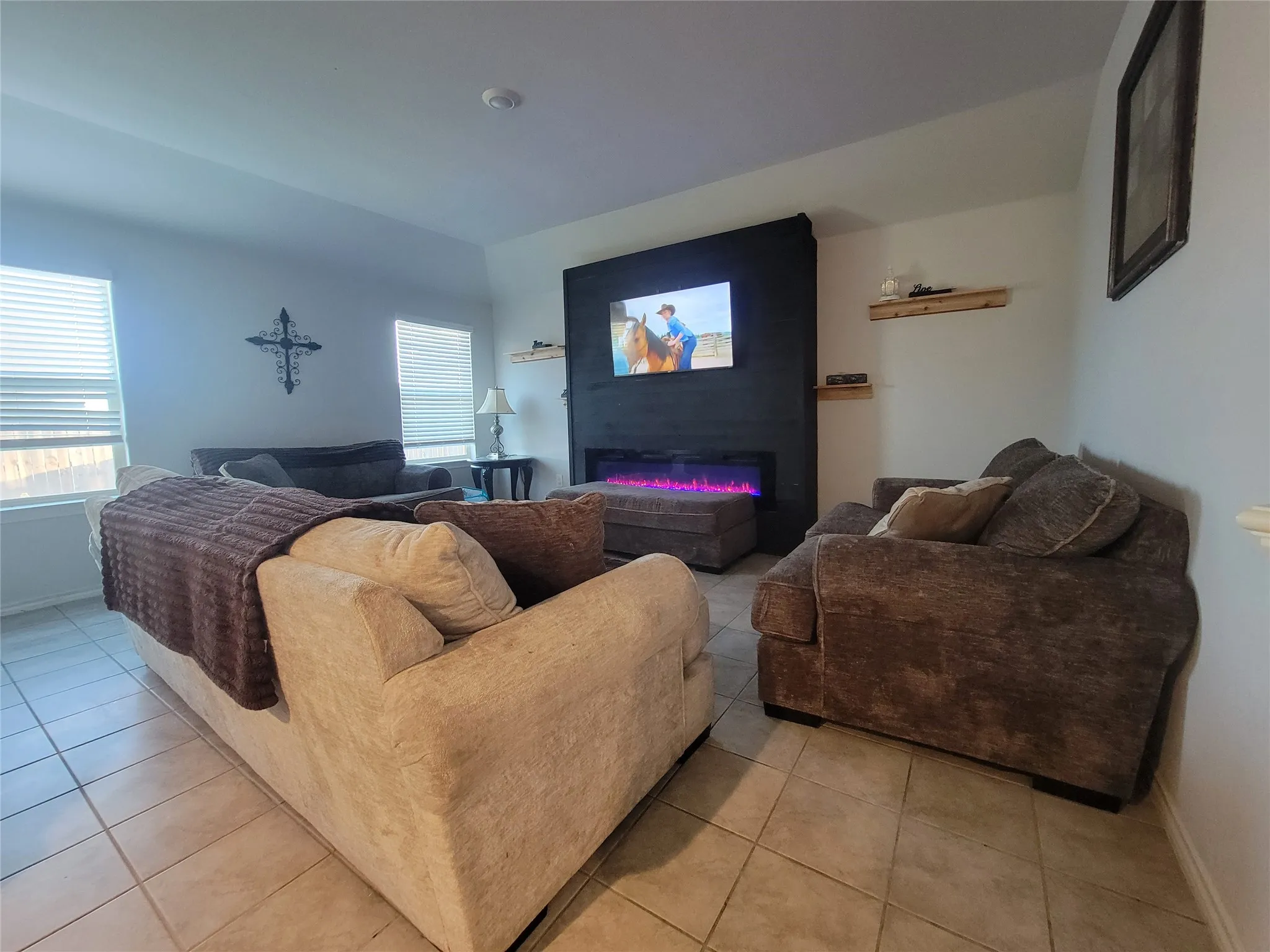 Tile flooring, white walls, two windows with blinds, a wall-mounted media console with an integrated fireplace, and two floating shelves
