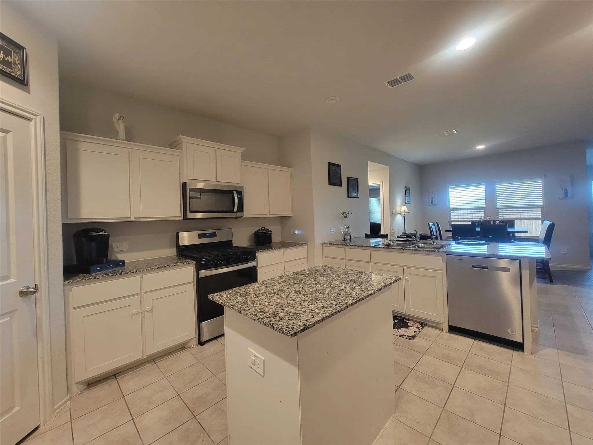 Open-concept kitchen featuring white cabinetry, speckled quartz countertops, a central island with seating space, stainless steel appliances including a microwave and dishwasher, tile flooring, and abundant natural light from multiple windows overlooking the adjacent living area