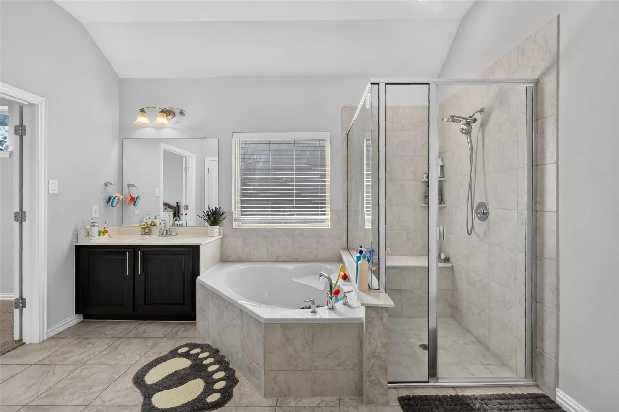 Alcove soaking tub, separate glass-enclosed shower, single vanity with dark cabinetry, wall-mounted light fixture, and tiled flooring