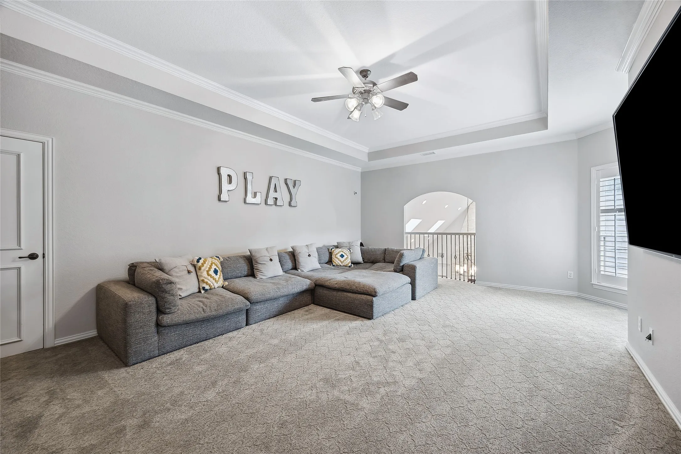 Spacious room featuring tray ceiling with crown molding, ceiling fan, archway to a landing, and window with plantation shutters