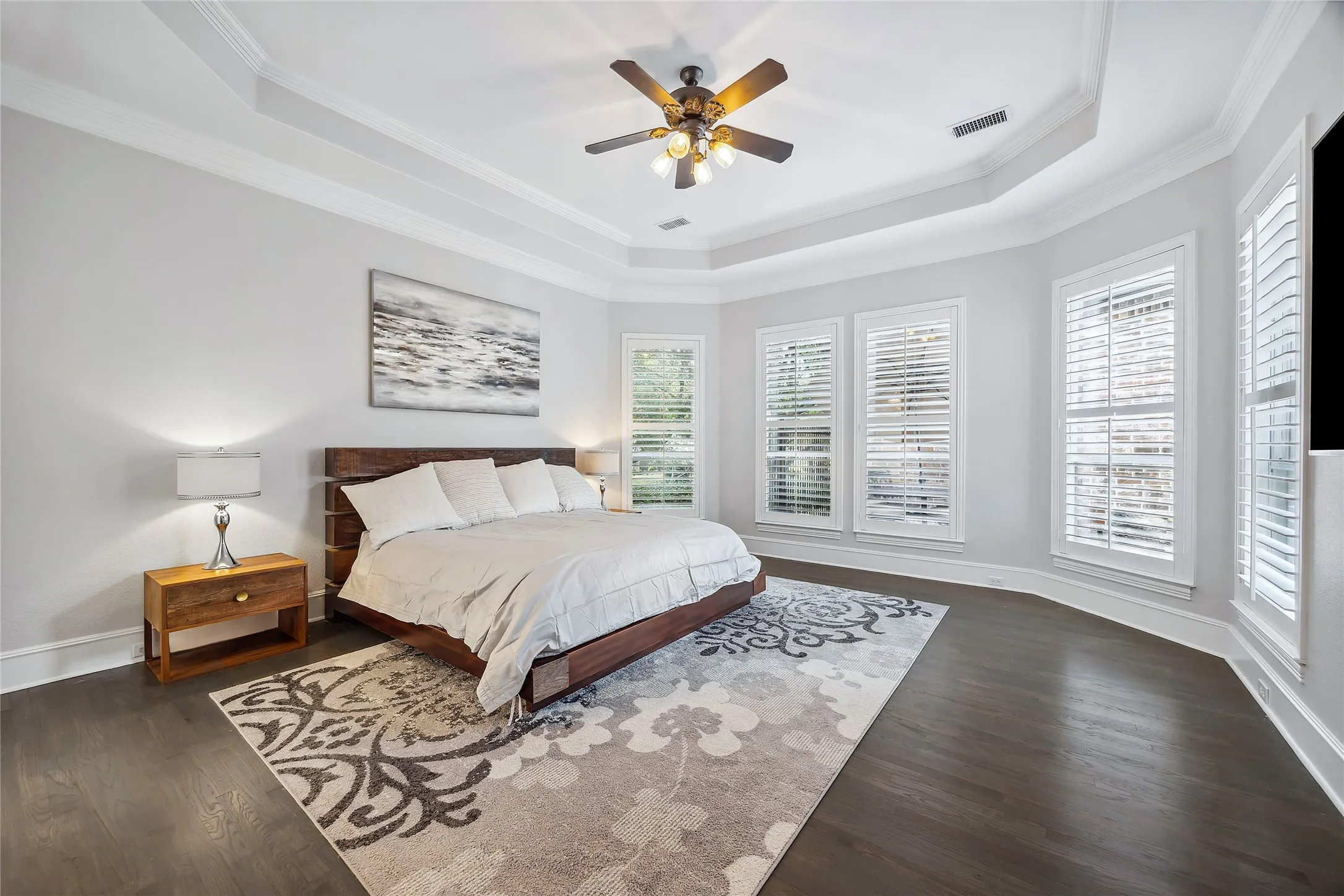 Tray ceiling with crown molding, ceiling fan, wood-finish flooring, multiple windows with white plantation shutters, and light gray wall paint