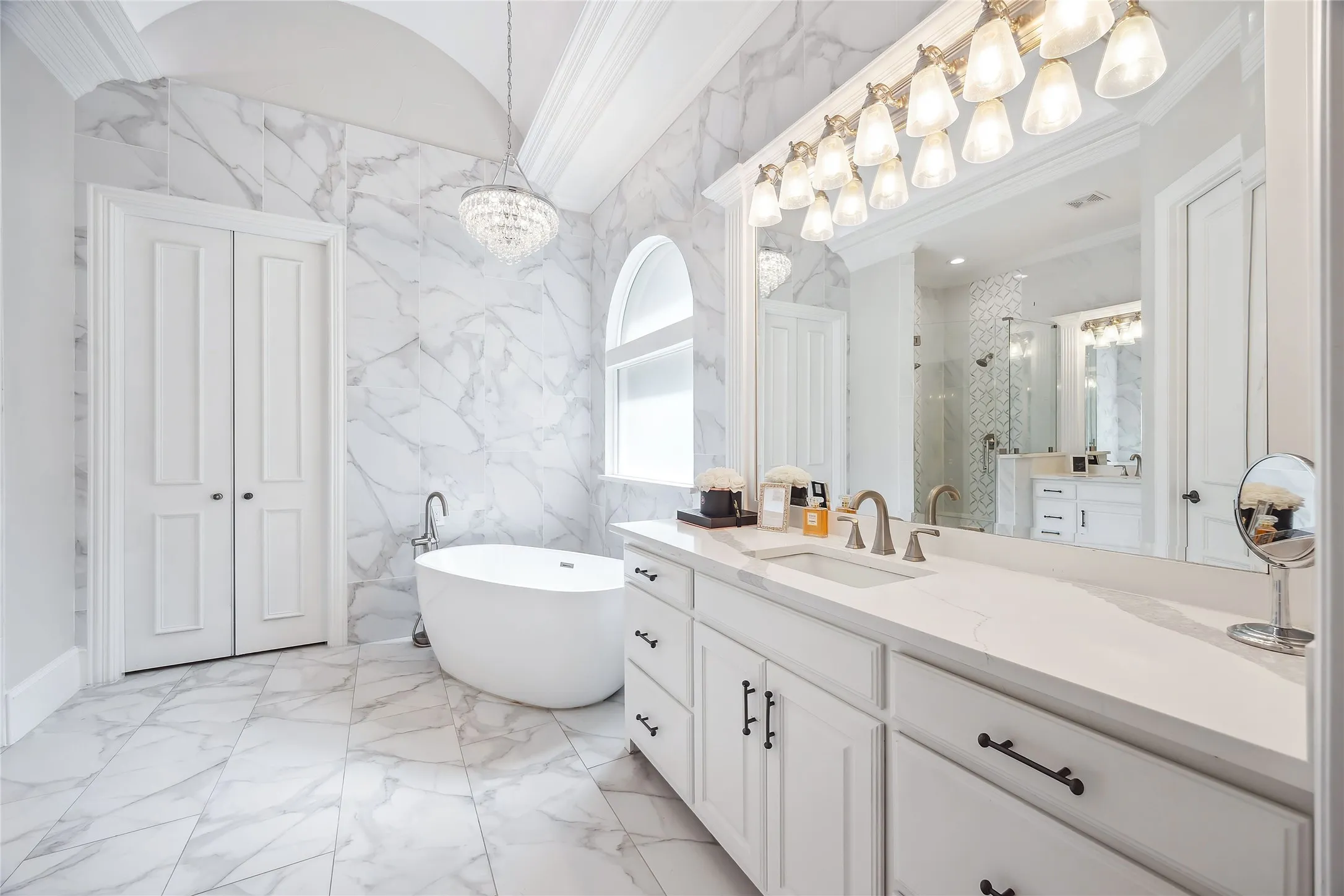 Freestanding soaking tub, floor-to-ceiling marbled tile, vaulted ceiling with chandelier, arched window, and dual vanity with integrated sink