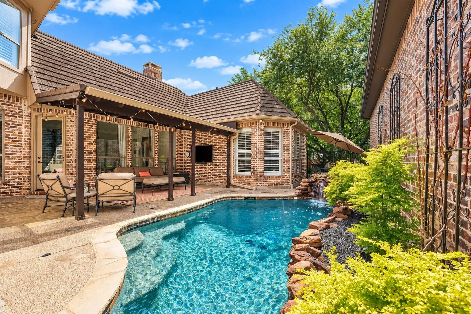 Private swimming pool with integrated waterfall feature, brick exterior, covered patio, wood-look roofing, and mature landscaping