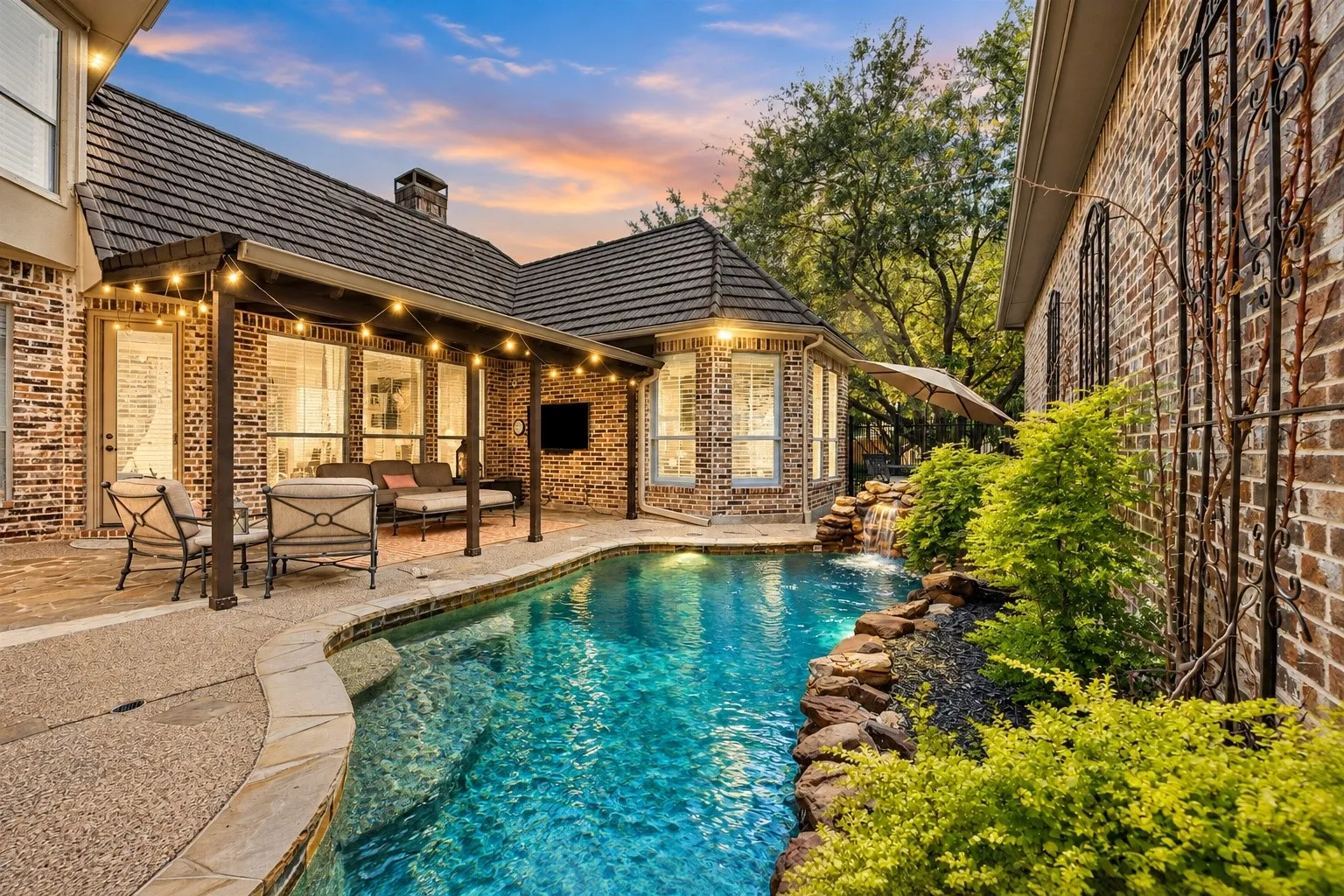 Brick exterior featuring a covered patio, a swimming pool with a stone waterfall, mature landscaping, and an ornate iron trellis