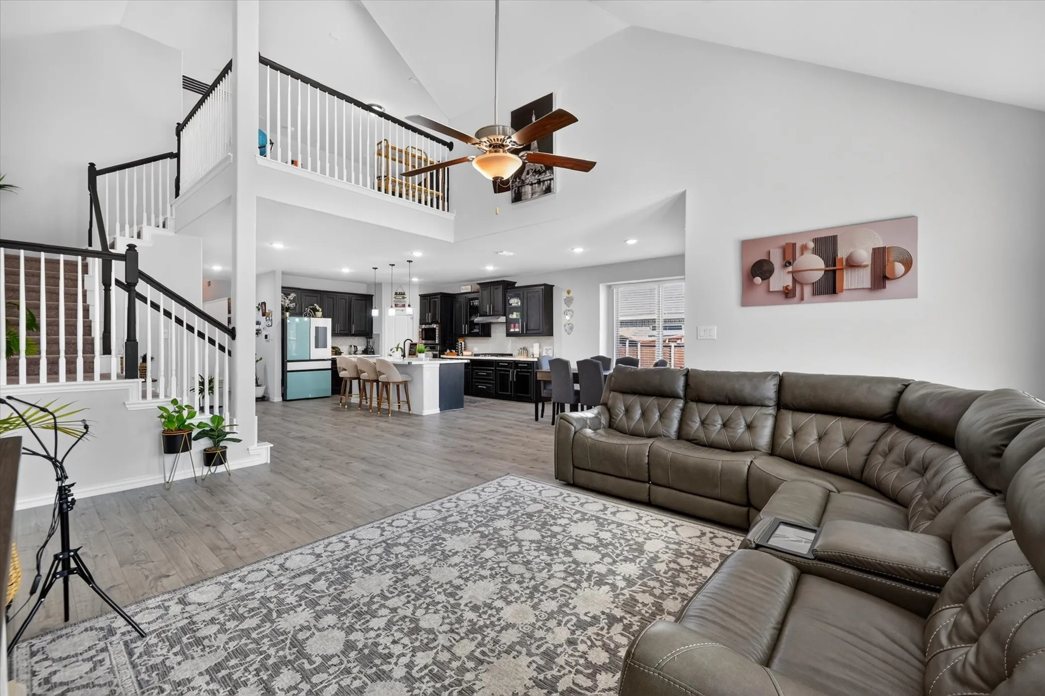 Vaulted ceiling with ceiling fan, wood-finish flooring, two-story staircase with black and white railings, recessed lighting, and a kitchen featuring a center island with seating