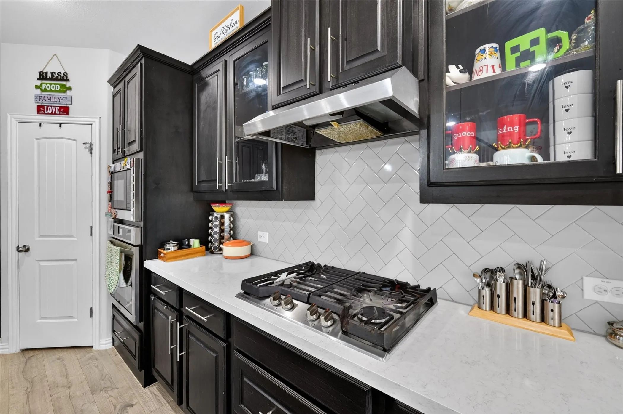 Dark wood-finish cabinetry with glass-front accents, stainless steel gas range, built-in microwave and oven, light-toned countertops, and herringbone tile​​‌​​​​‌​​‌‌​‌‌​​​‌‌​‌​‌​‌​​​‌​​ backsplash