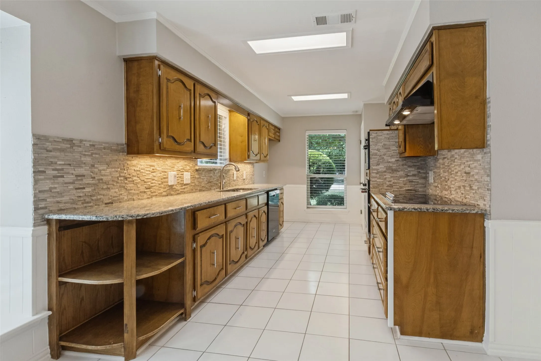Wood cabinetry with decorative door panels, granite-style countertops, mosaic tile backsplash, under-cabinet lighting, and white tile flooring