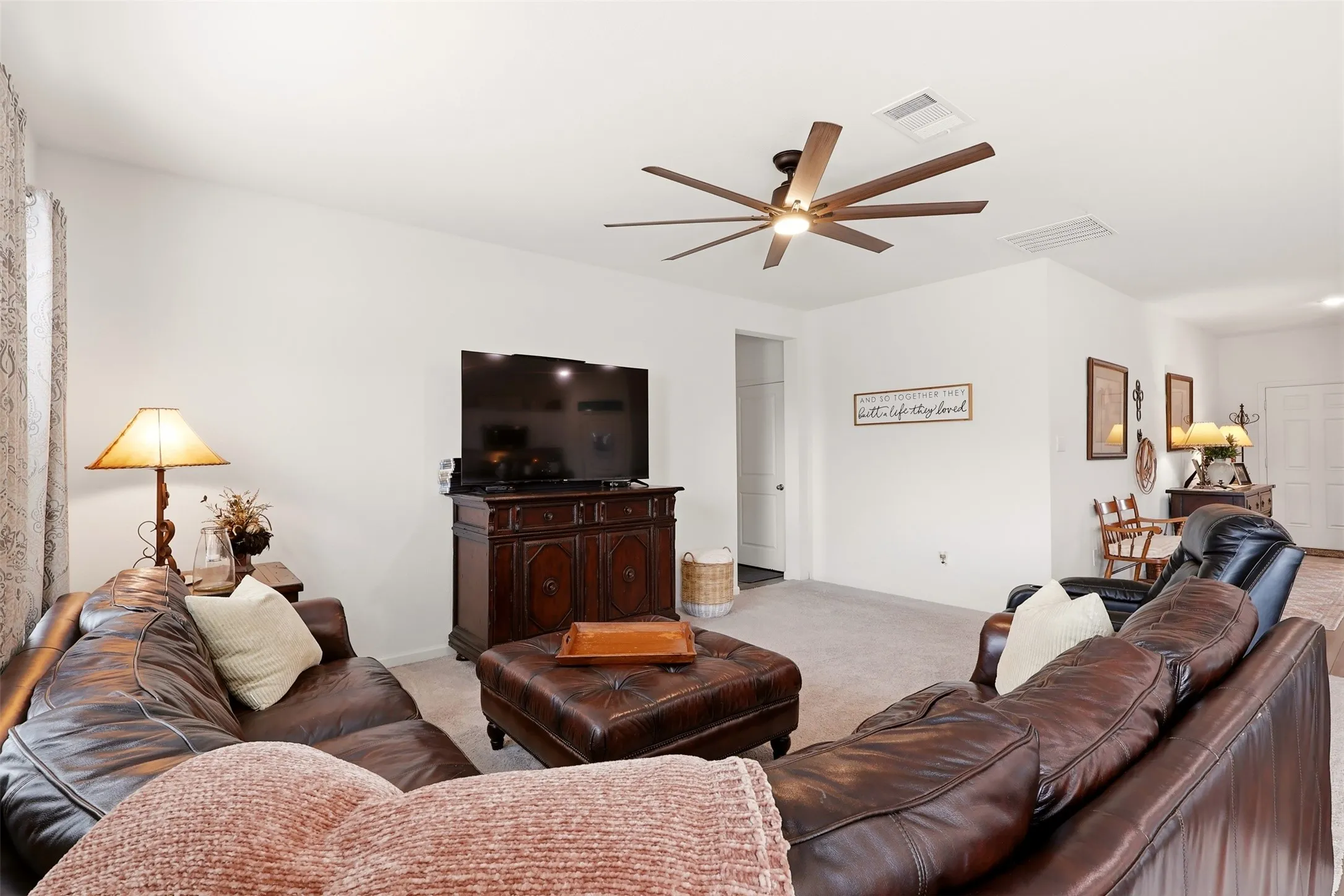 Spacious living area featuring light-toned walls, ceiling fan with integrated lighting, and carpeted flooring