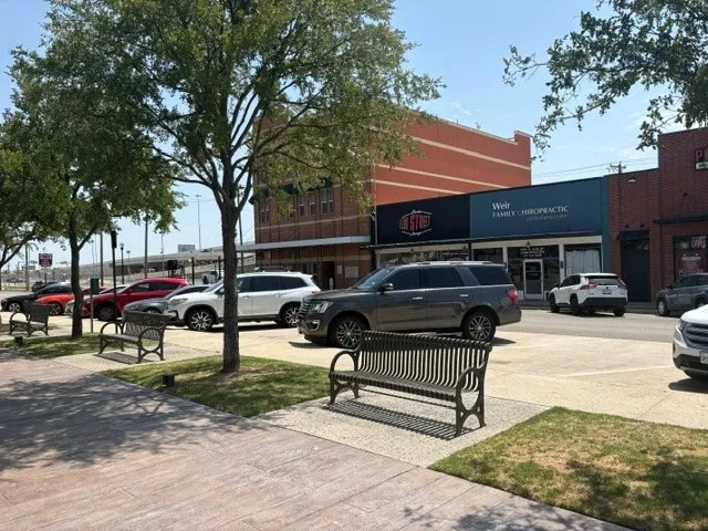 Sidewalk area featuring mature trees, landscaped planting beds, and multiple public benches