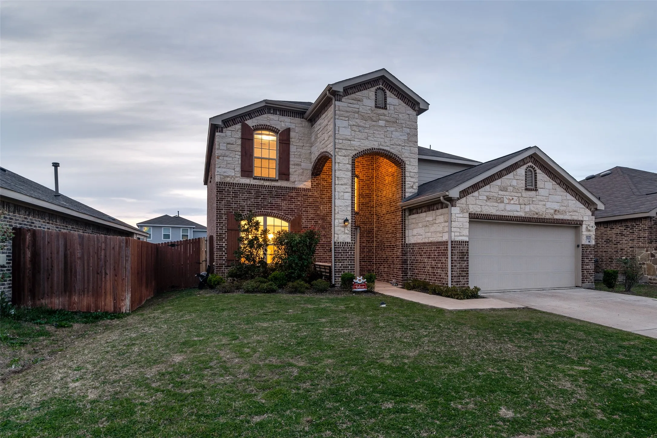 Brick and stone facade, arched entryway, attached two-car garage, mature landscaping, and privacy fencing