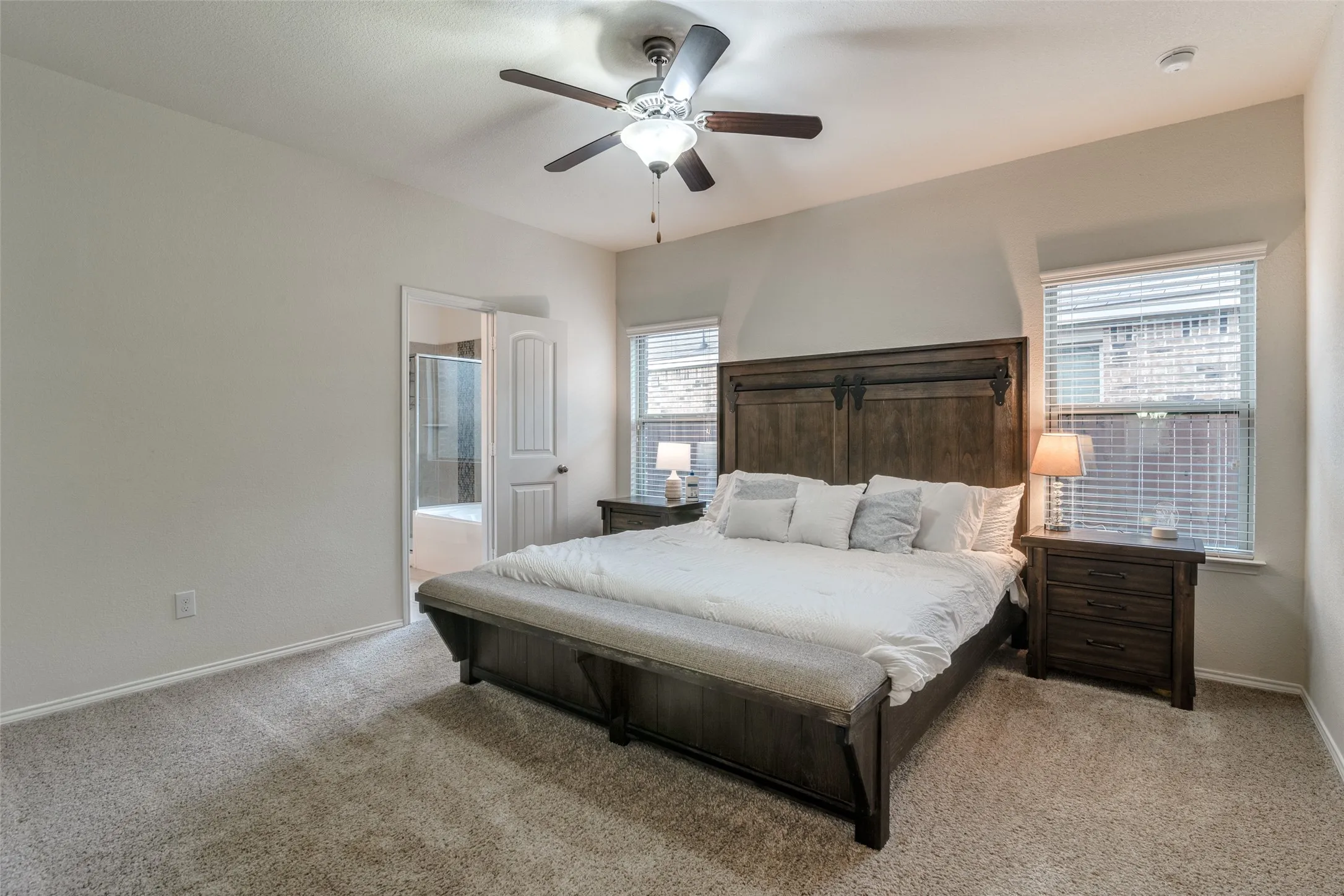 Carpeted bedroom with a ceiling fan, two windows with blinds, and an attached bathroom featuring a shower-tub combination
