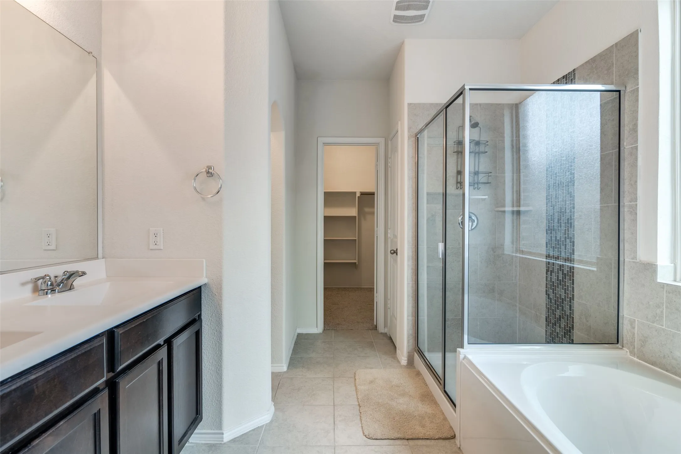 Dual vanity with dark wood-finish cabinetry, tile flooring, and a glass-enclosed shower over a built-in tub