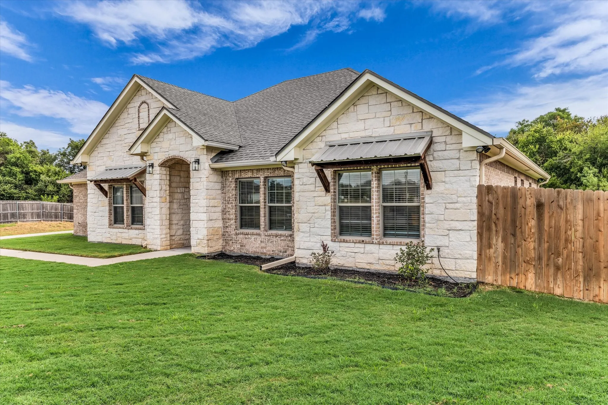 Stone and brick exterior with a shingle roof, arched entry, decorative window awnings, and a wood privacy fence