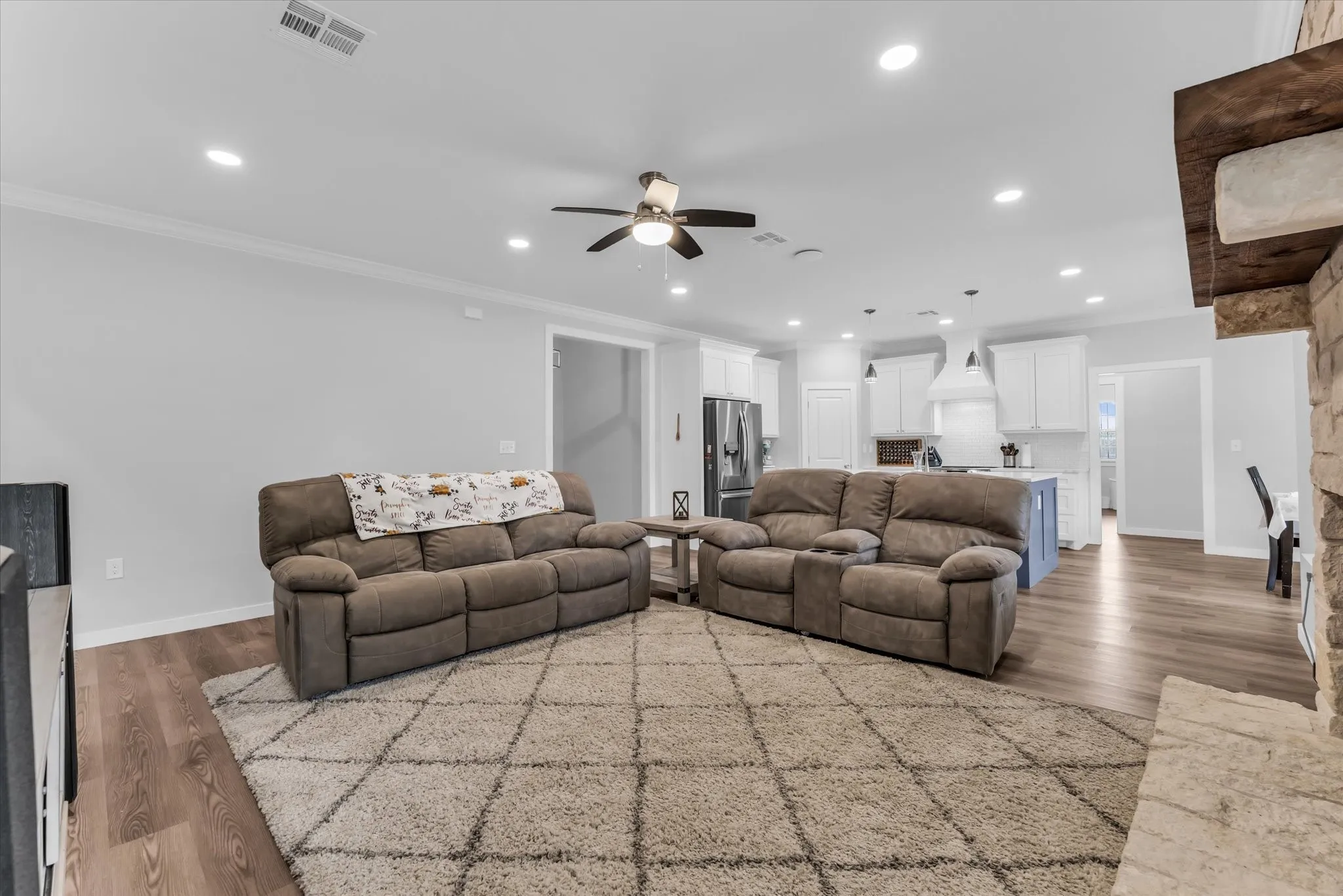 Open concept living space featuring recessed lighting, a ceiling fan, crown molding, wood-finish flooring, and a stone fireplace surround with a wood mantel