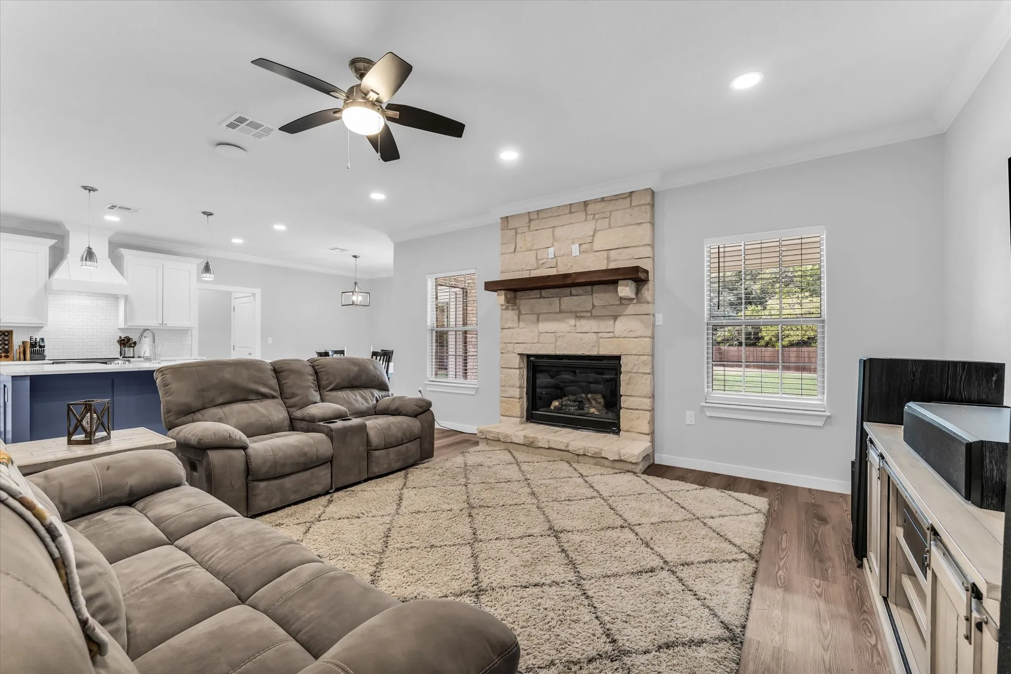 Stacked stone fireplace with a wood mantel, overhead recessed lighting, wood-finish flooring, and a ceiling fan with integrated lighting