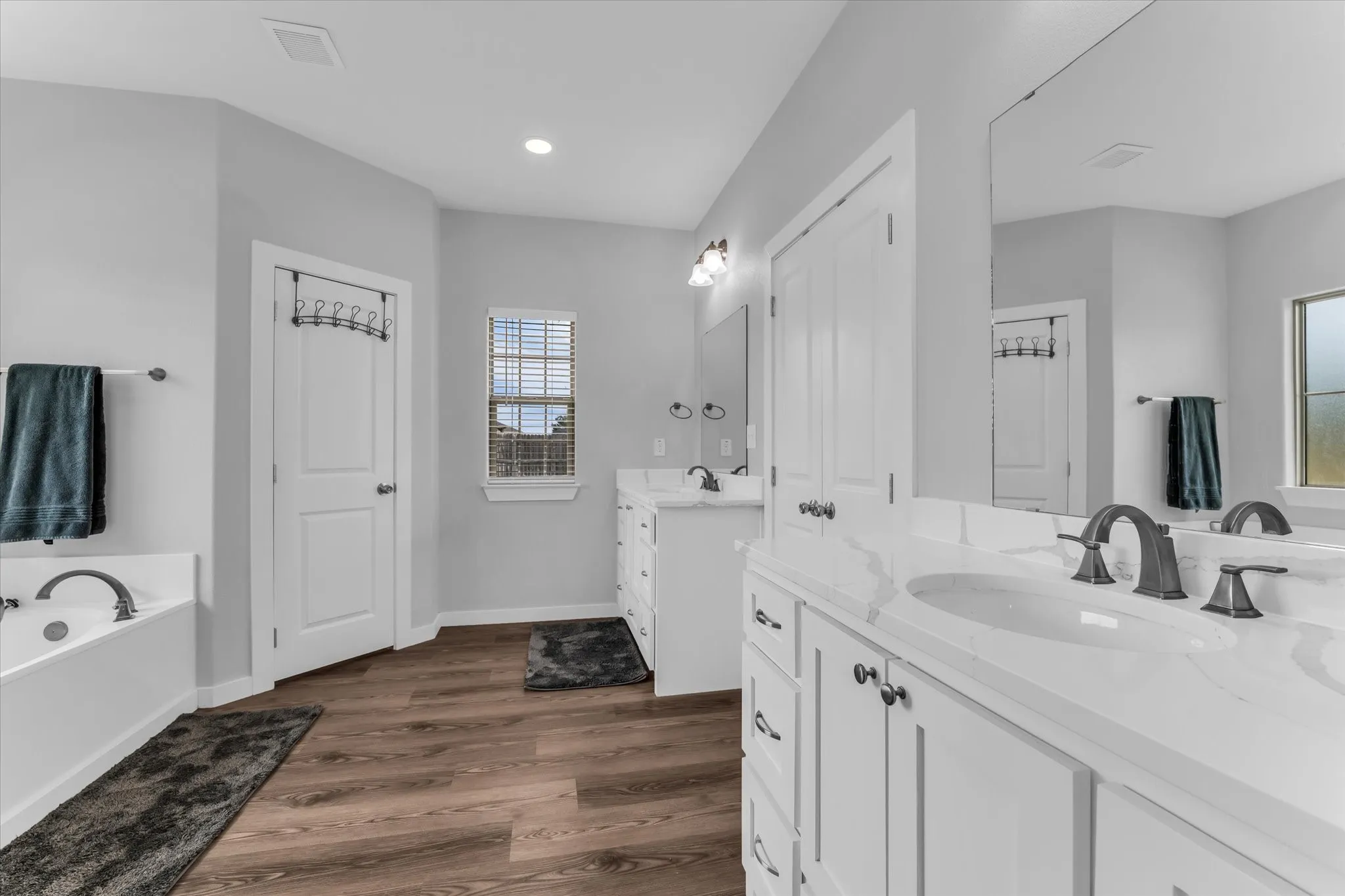 White double vanity with integrated sinks and dark bronze fixtures, a built-in soaking tub, a window with horizontal blinds, and wood-finish flooring