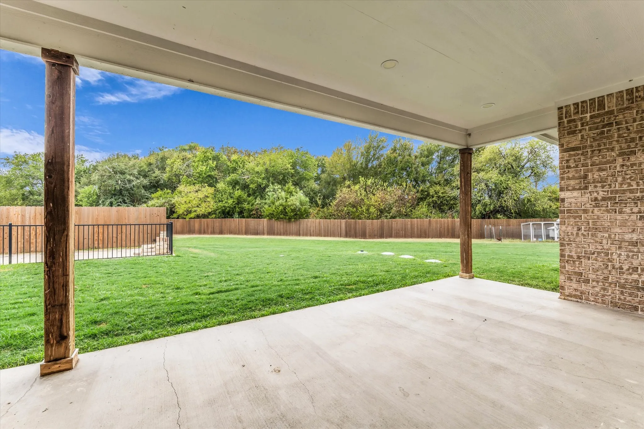 Covered concrete patio with recessed lighting, supported by wood-finish pillars, overlooking a fenced backyard with a manicured lawn and mature trees